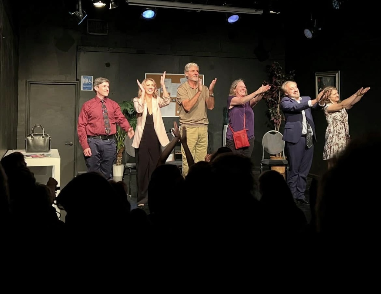 Six people stand on stage, clapping and smiling, with audience silhouettes in foreground, black background, stage lights above.