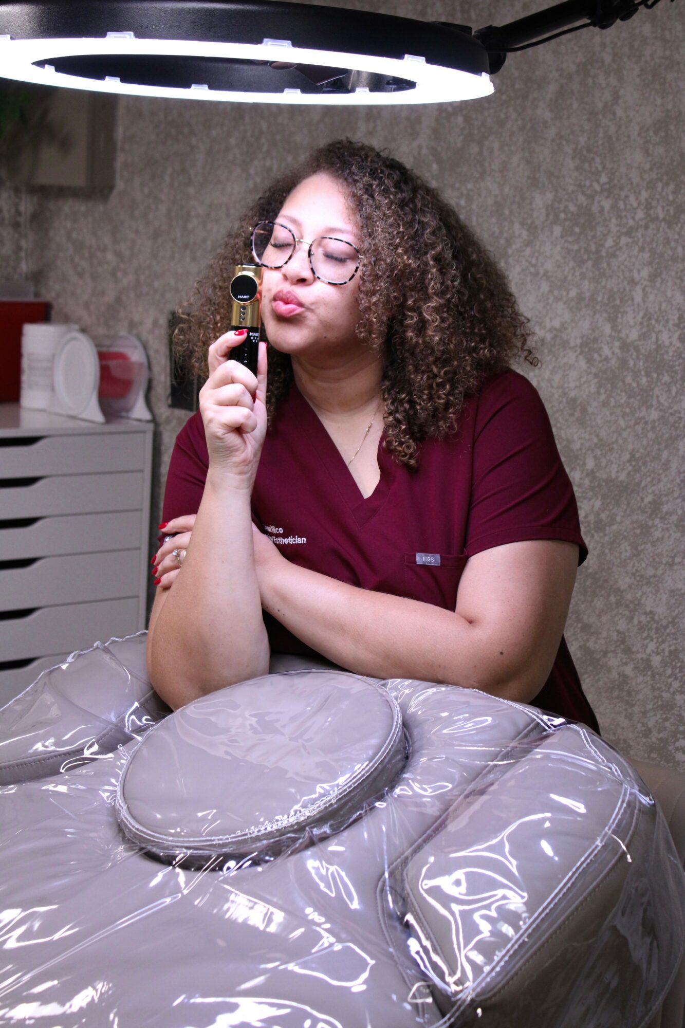 Woman with curly hair and glasses sitting at table, holding a camera, with a ring light overhead.