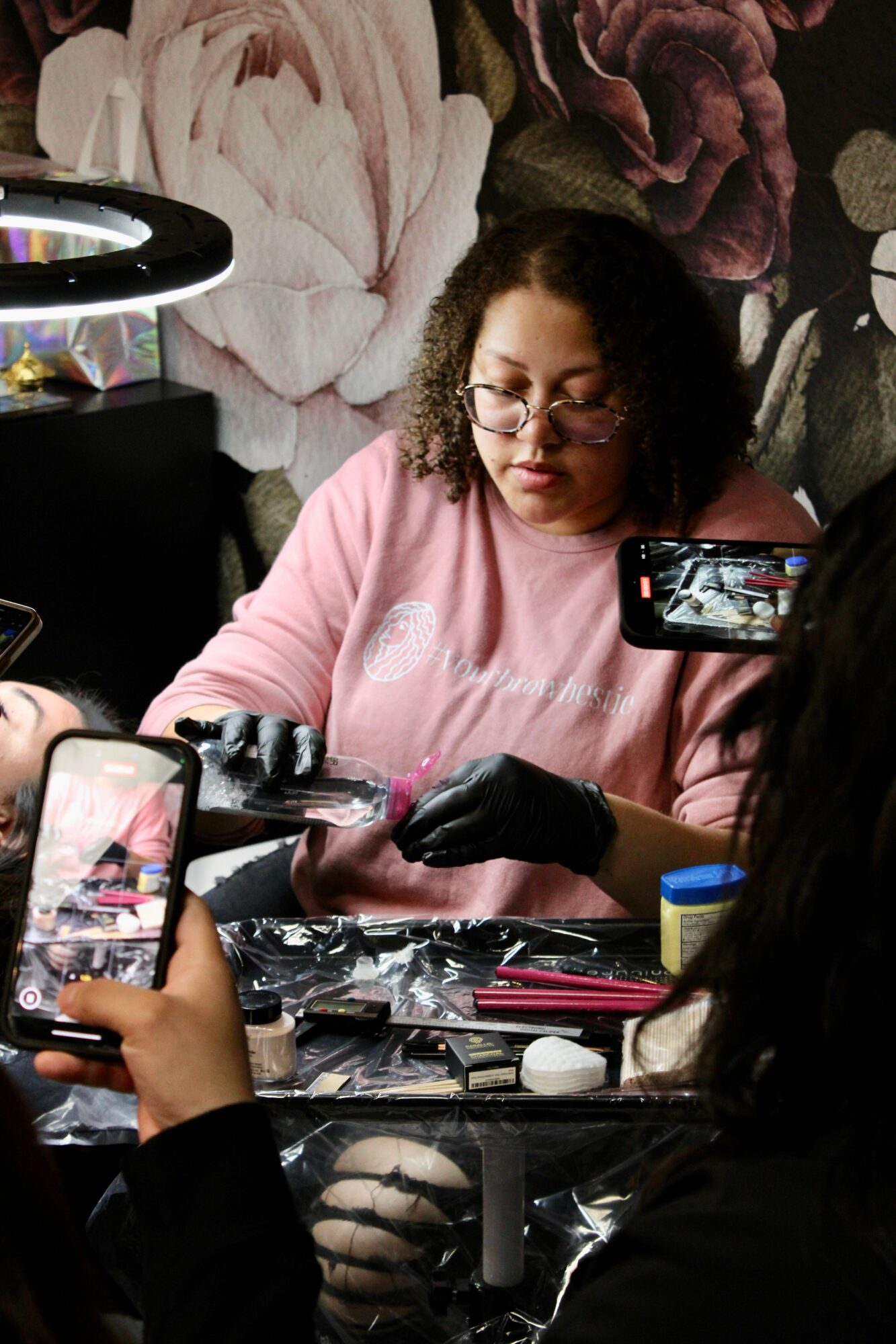 Woman with curly hair and glasses working on a tattoo design, surrounded by tattoo equipment and people taking photos.