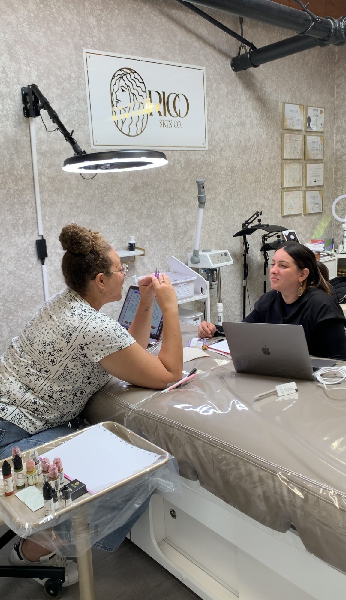 Two women sitting at a table in a nail salon, one with a laptop, the other with a nail file, engaging in conversation.