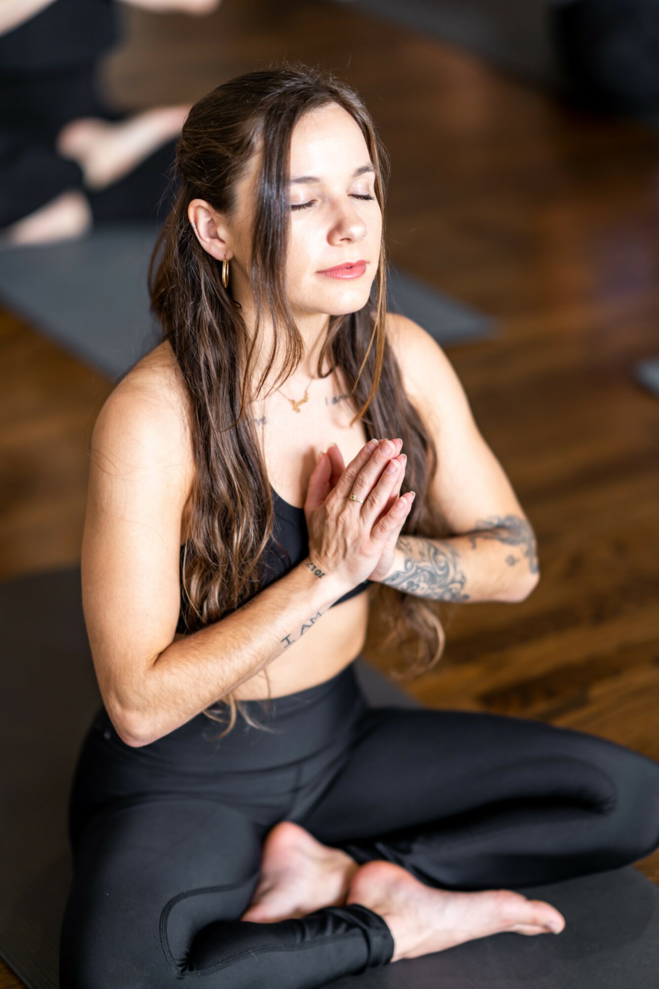 Woman with long dark hair sitting cross-legged with hands in prayer position, eyes closed, on wooden floor.