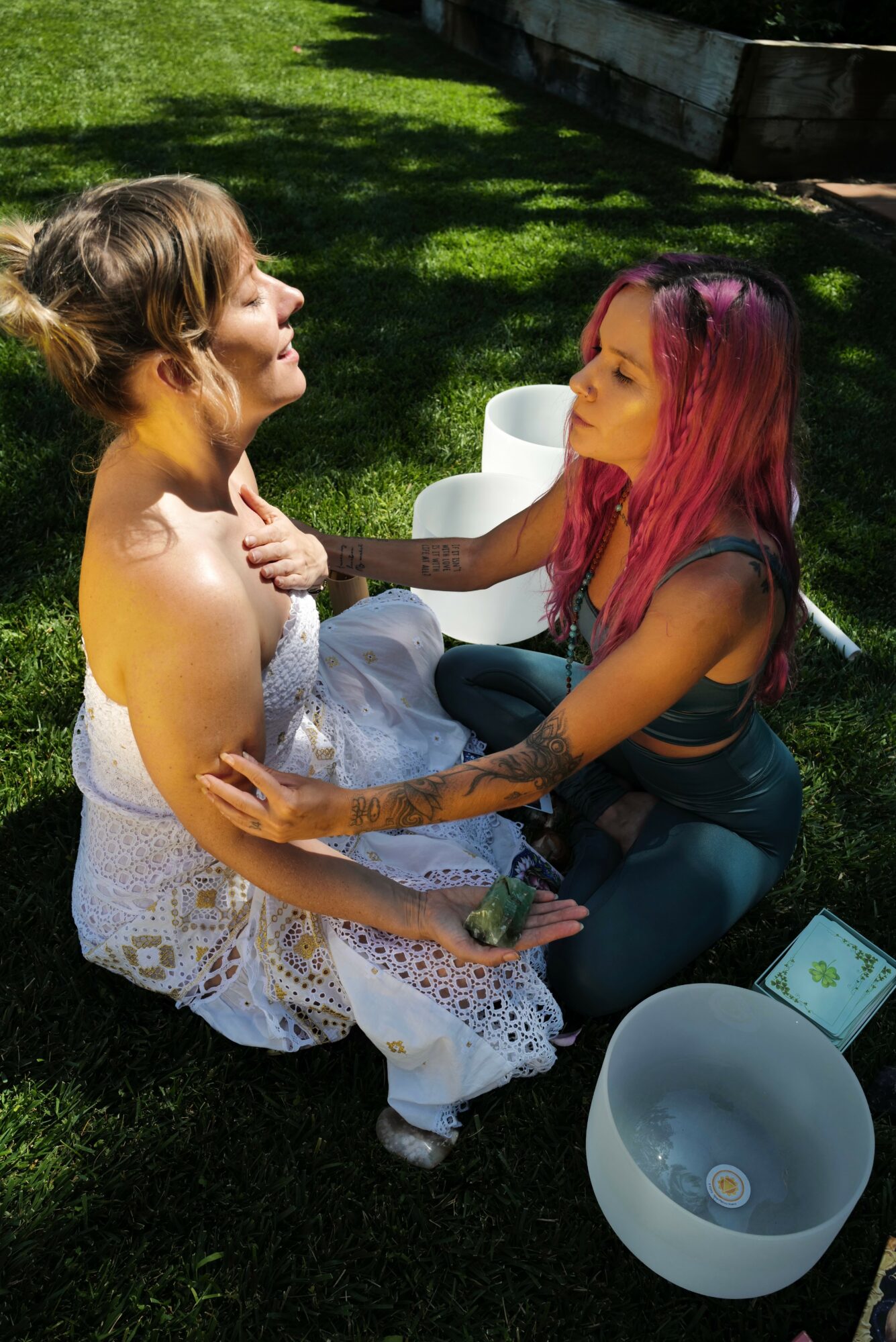 Two women sit on grass, one with pink hair and the other with brown hair, engaging in a conversation outdoors.