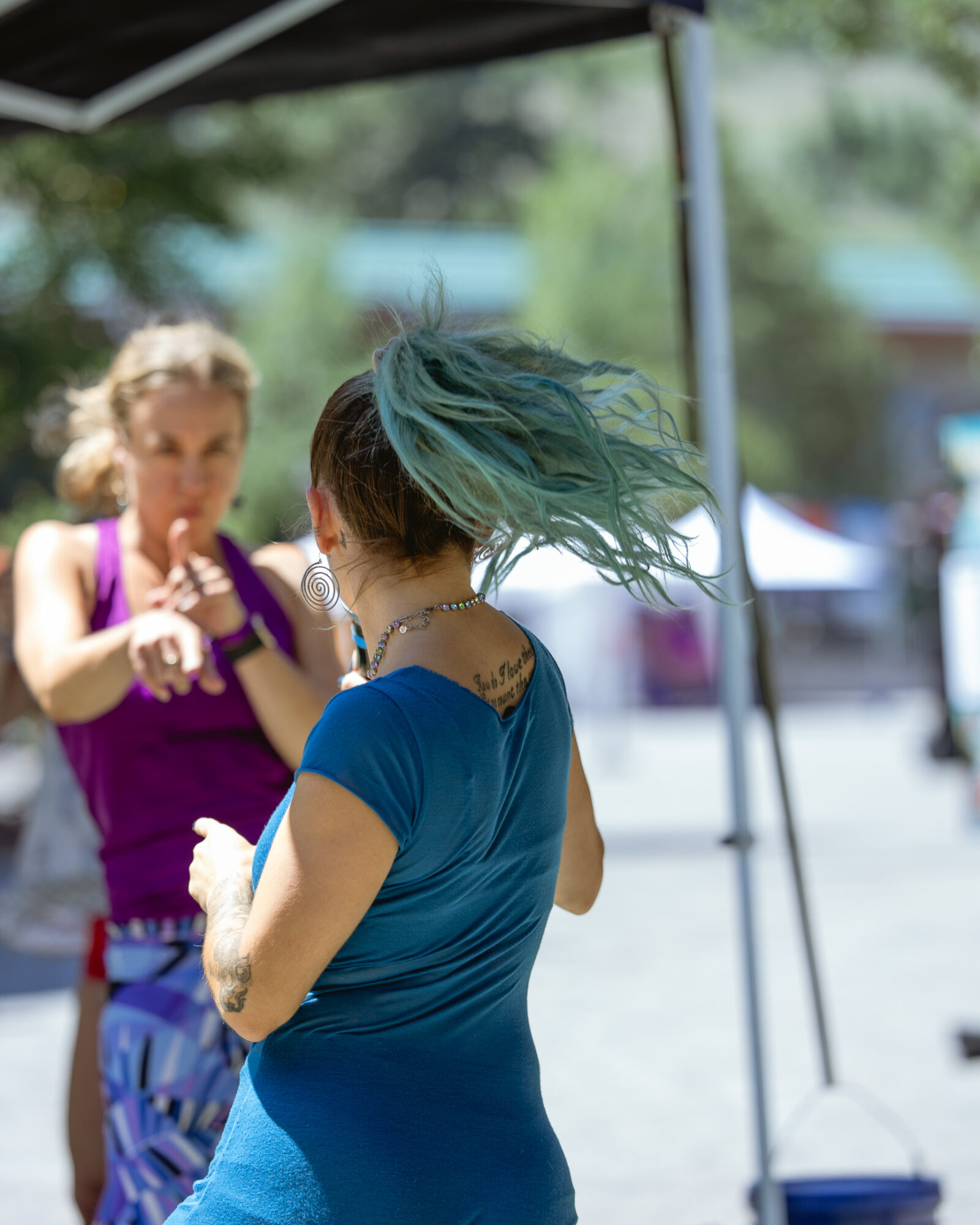 Person with teal hair and blue shirt standing outdoors, with another person in purple in the background.