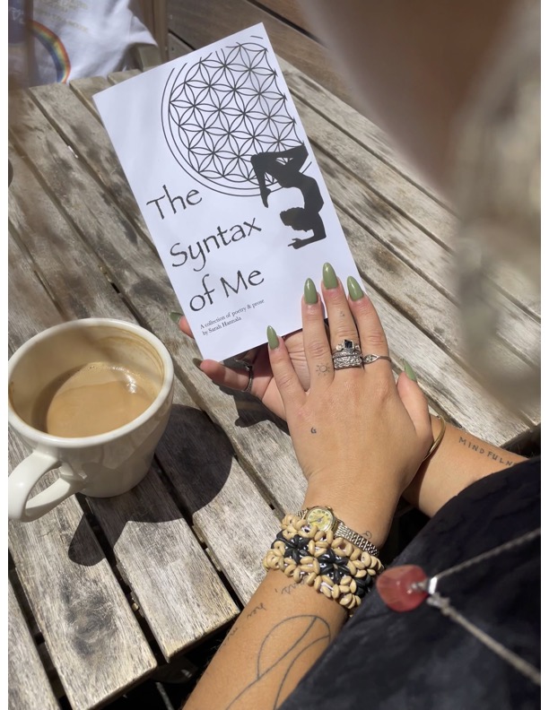 Person's hand with rings and bracelet, on wooden table with coffee cup and booklet titled 'The Syntax of Me'.