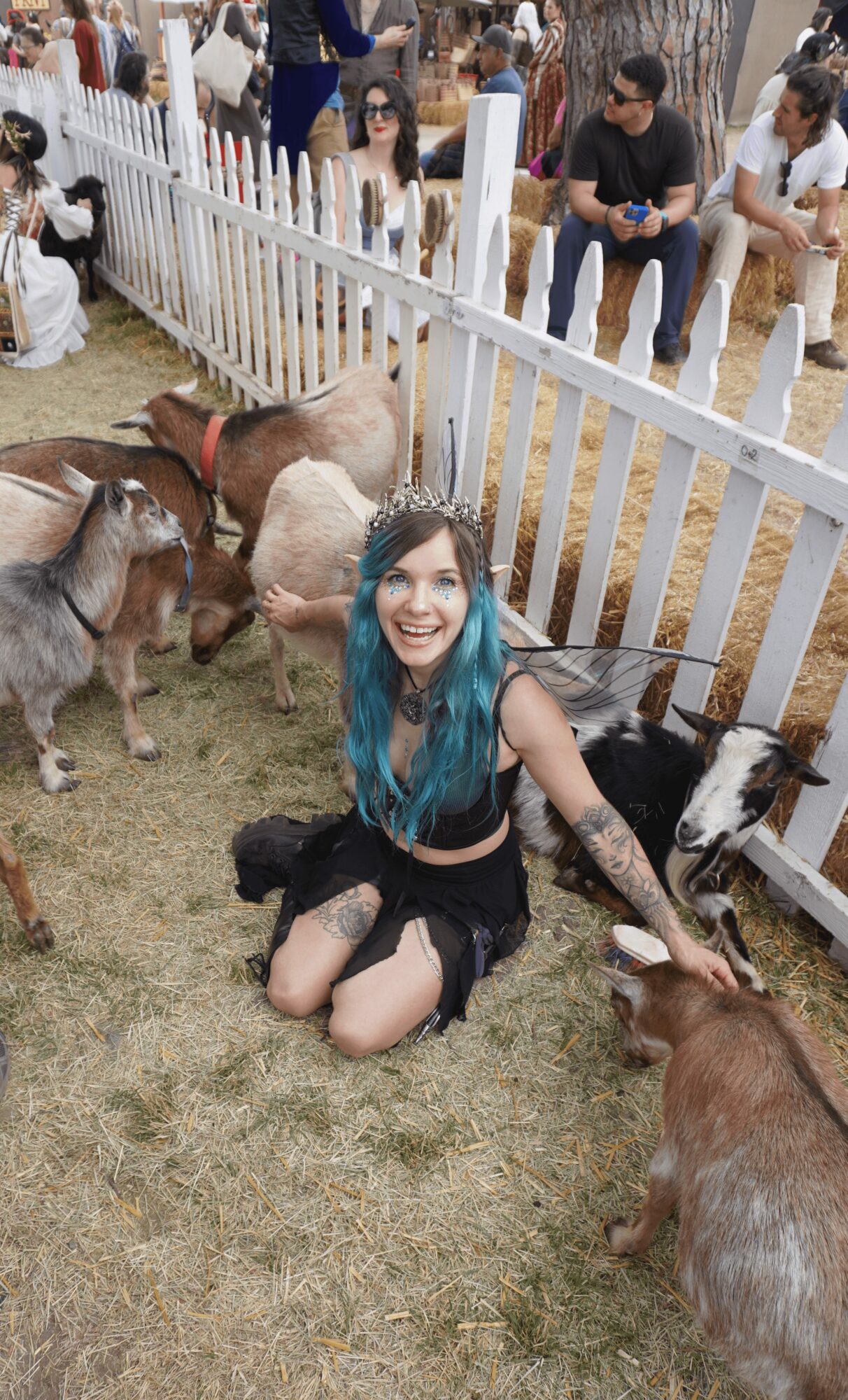 Young woman with blue hair kneeling on grass, petting goats near a white picket fence, with people in the background.