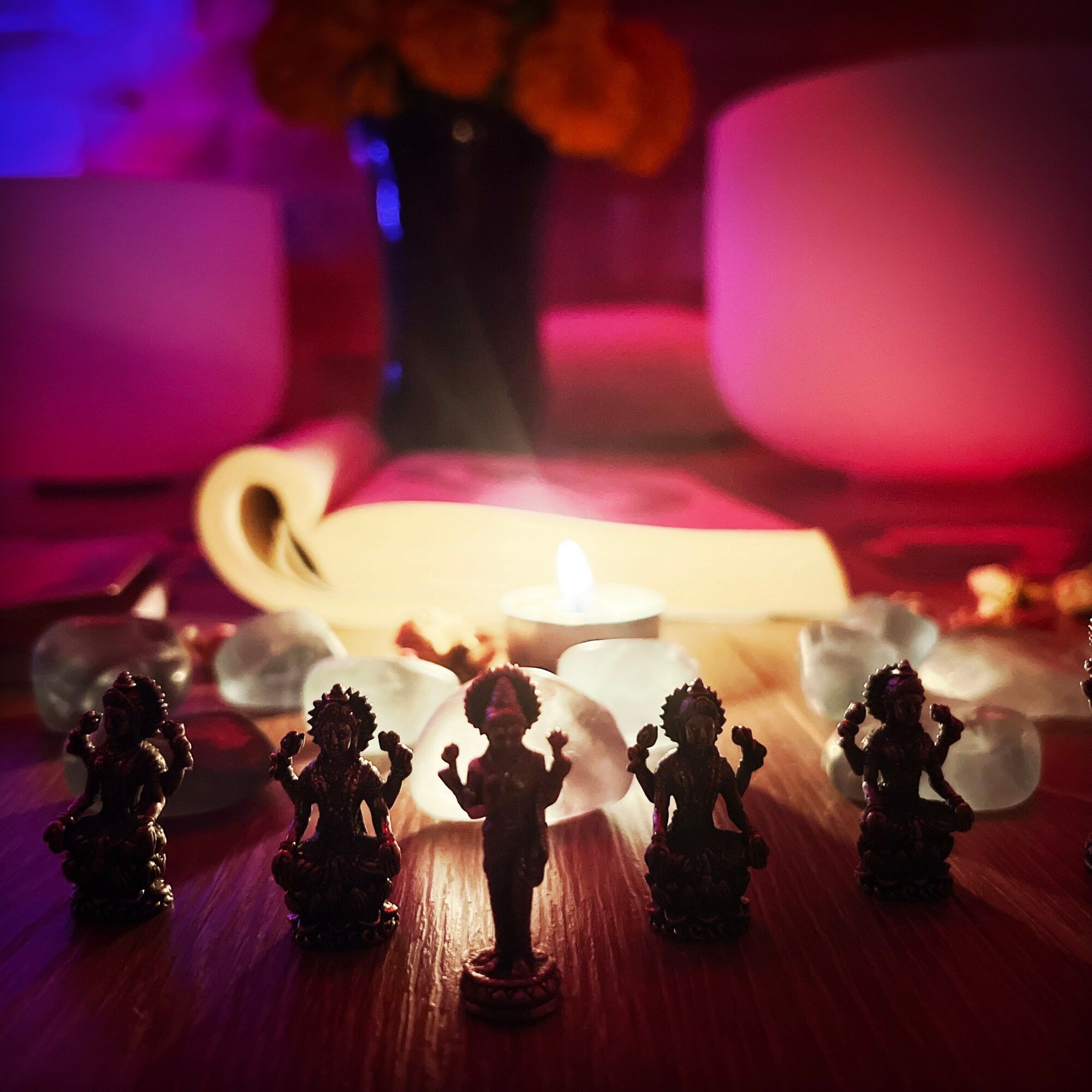 Silhouettes of five small statues in front of a lit candle, with blurred background of flowers and pink lighting.