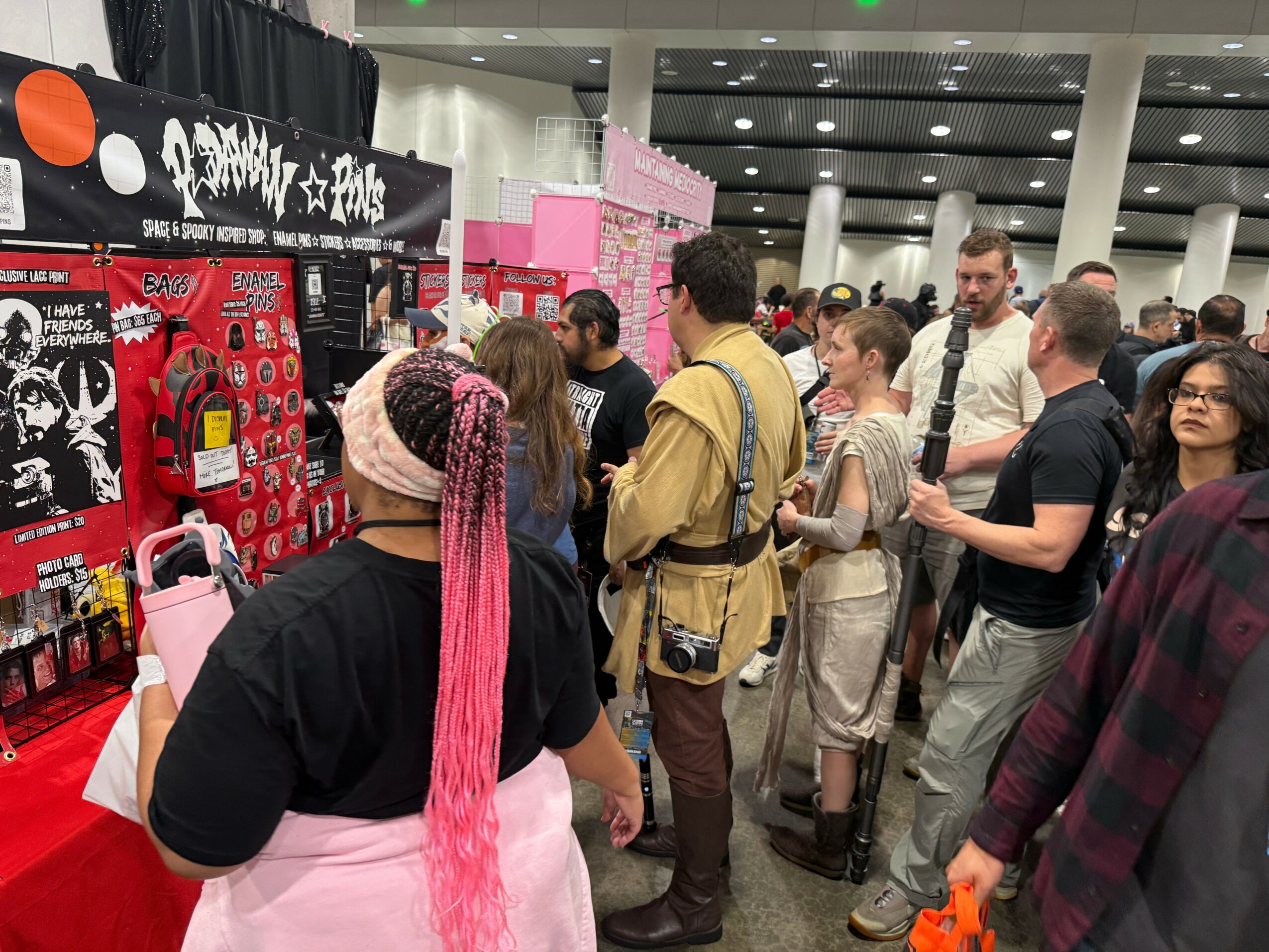 Crowd of people browsing booths at an indoor convention or market, with merchandise displays and banners visible.