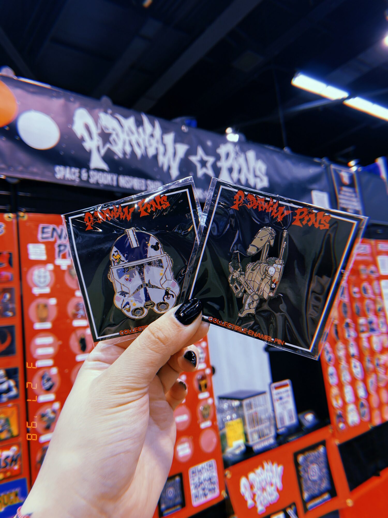 Person holding two square pins with artwork, in front of a red and black display at a convention or store.