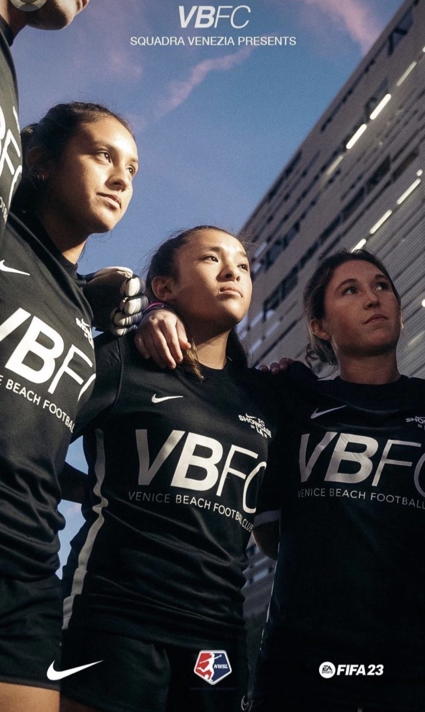 Three female soccer players in black jerseys standing outdoors with a building and blue sky behind them.