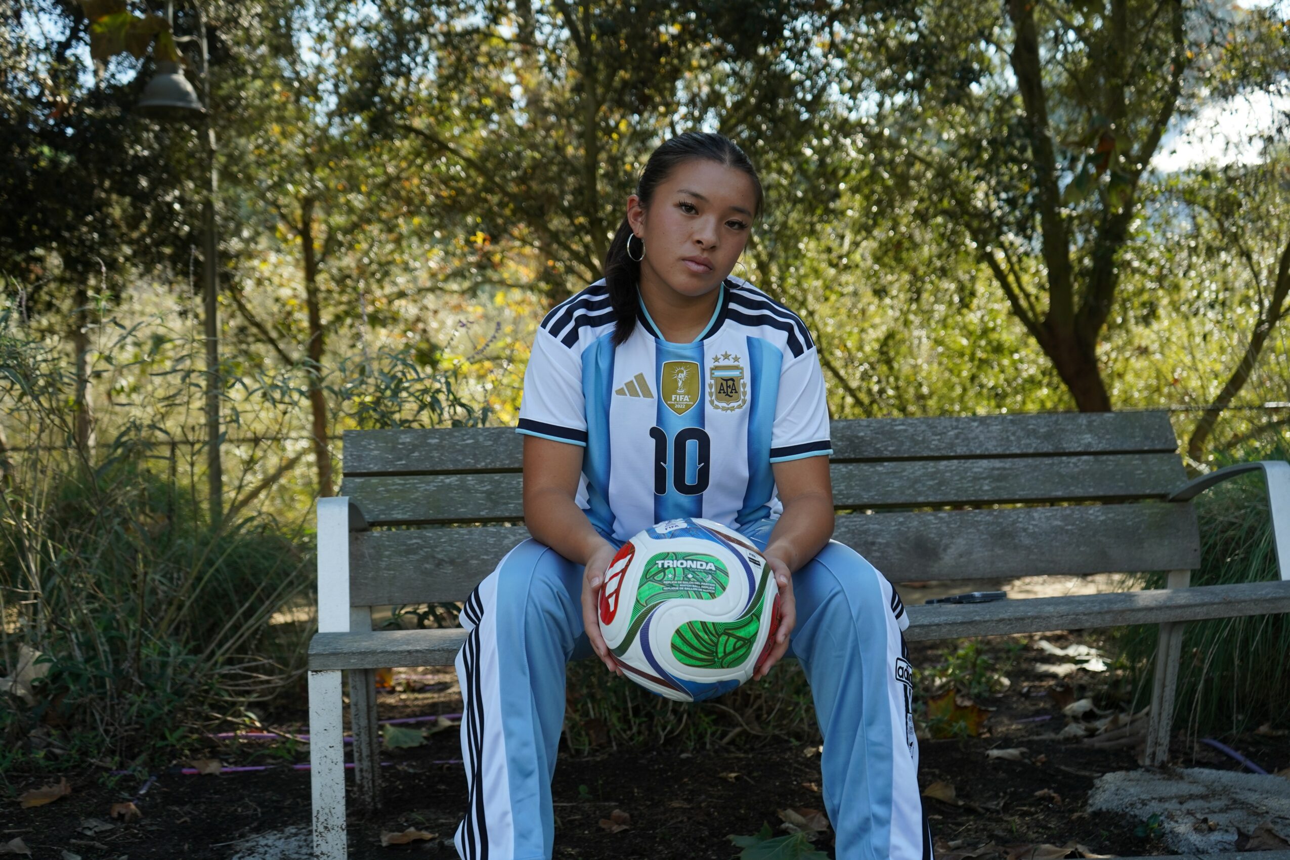 Female soccer player sitting on a bench holding a green and white soccer ball outdoors.