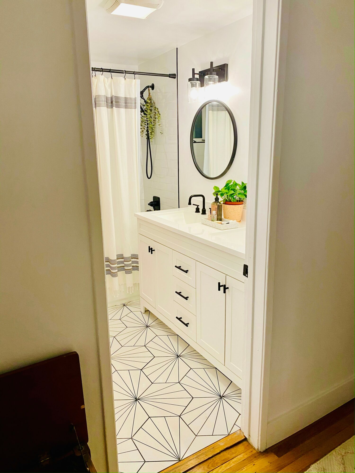 Bathroom with white vanity, oval mirror, potted plant, shower with curtain, geometric floor tiles, and wall shelf.