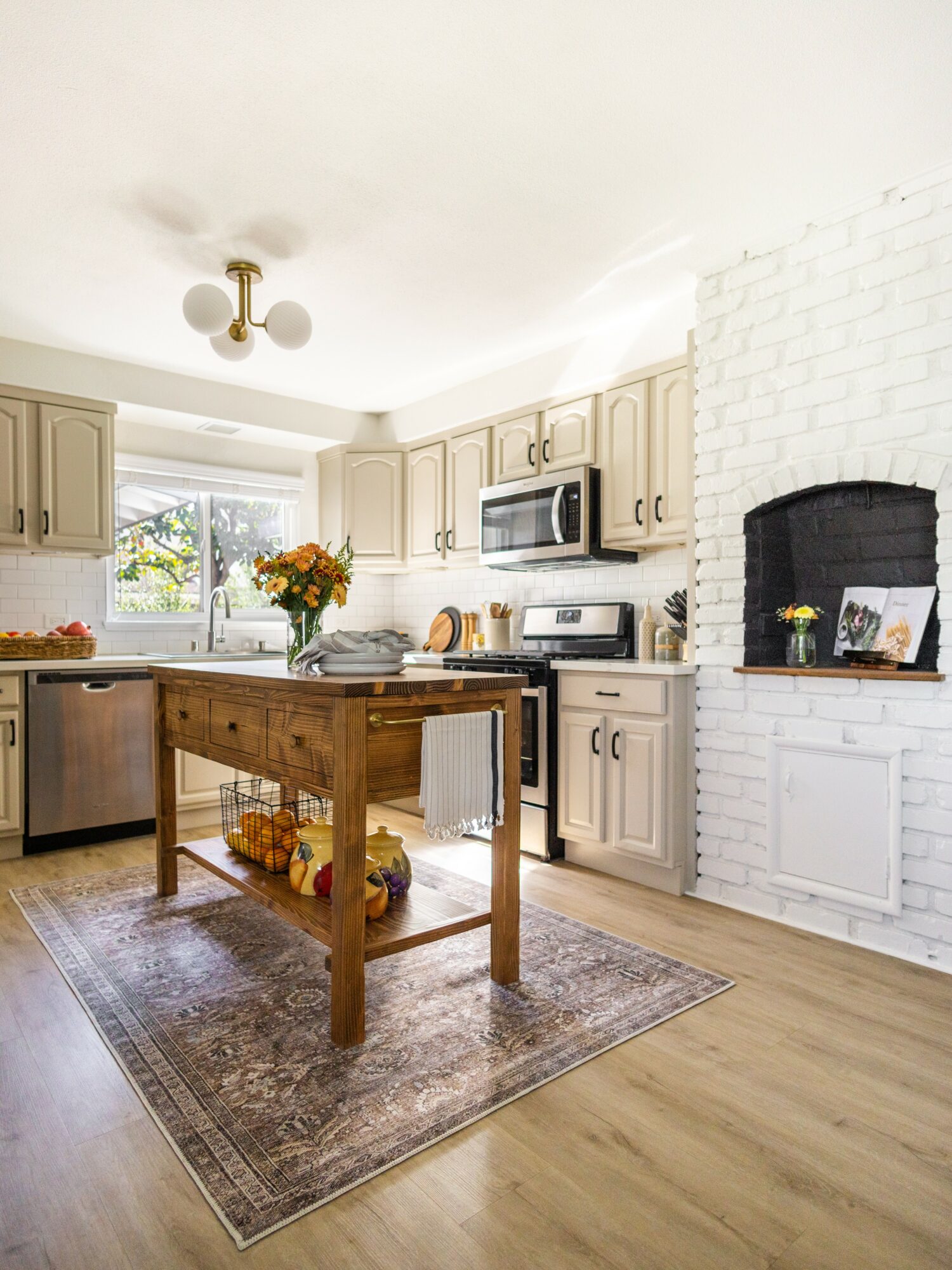 Kitchen with white cabinets, a wooden island, a window with a view, and a white brick fireplace with a black interior.