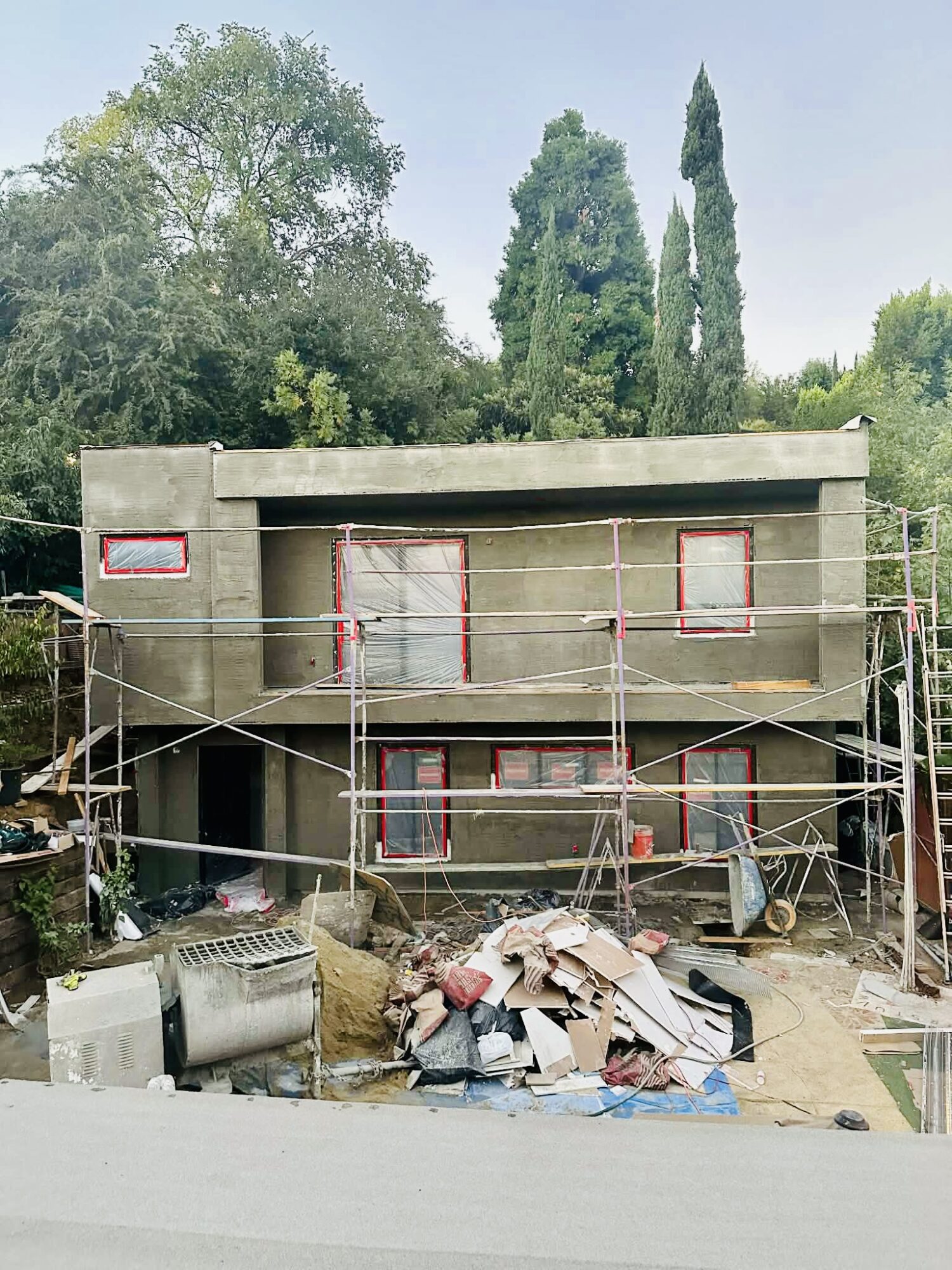 Three-story building under construction with scaffolding, surrounded by trees and construction debris.