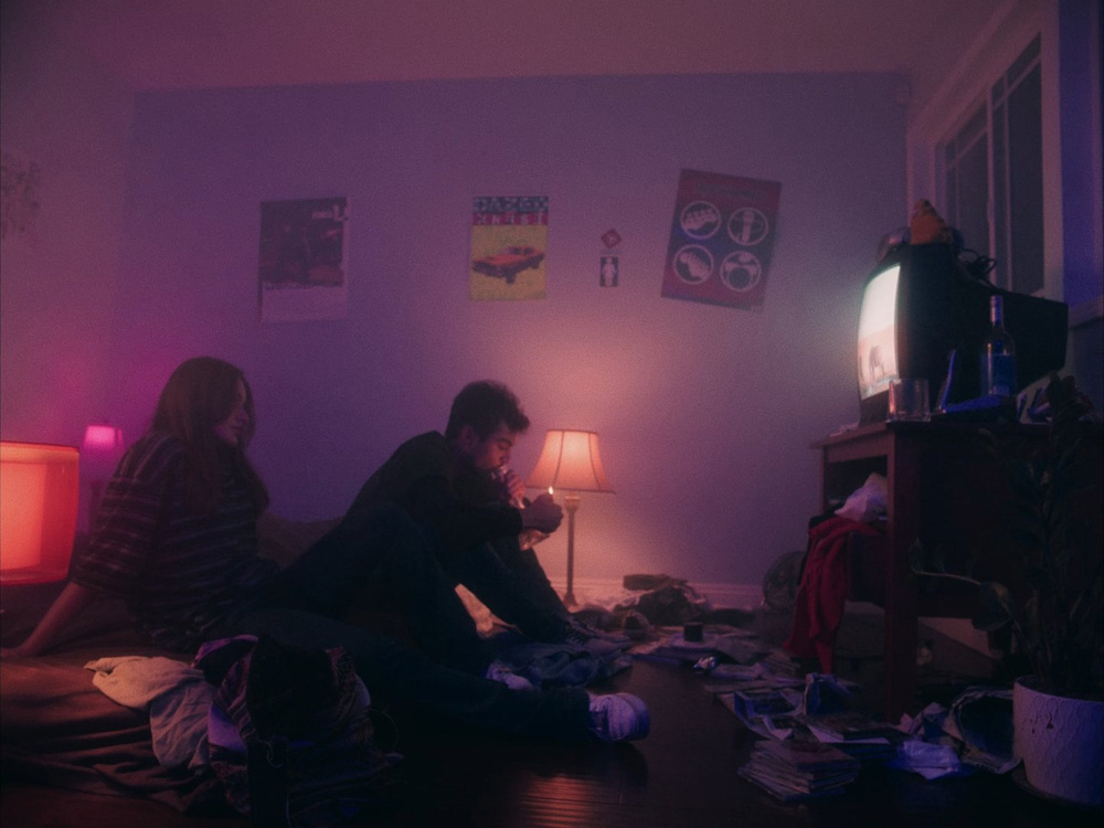 Two people sit on a bed in a dimly lit room watching television with cluttered items on the floor.