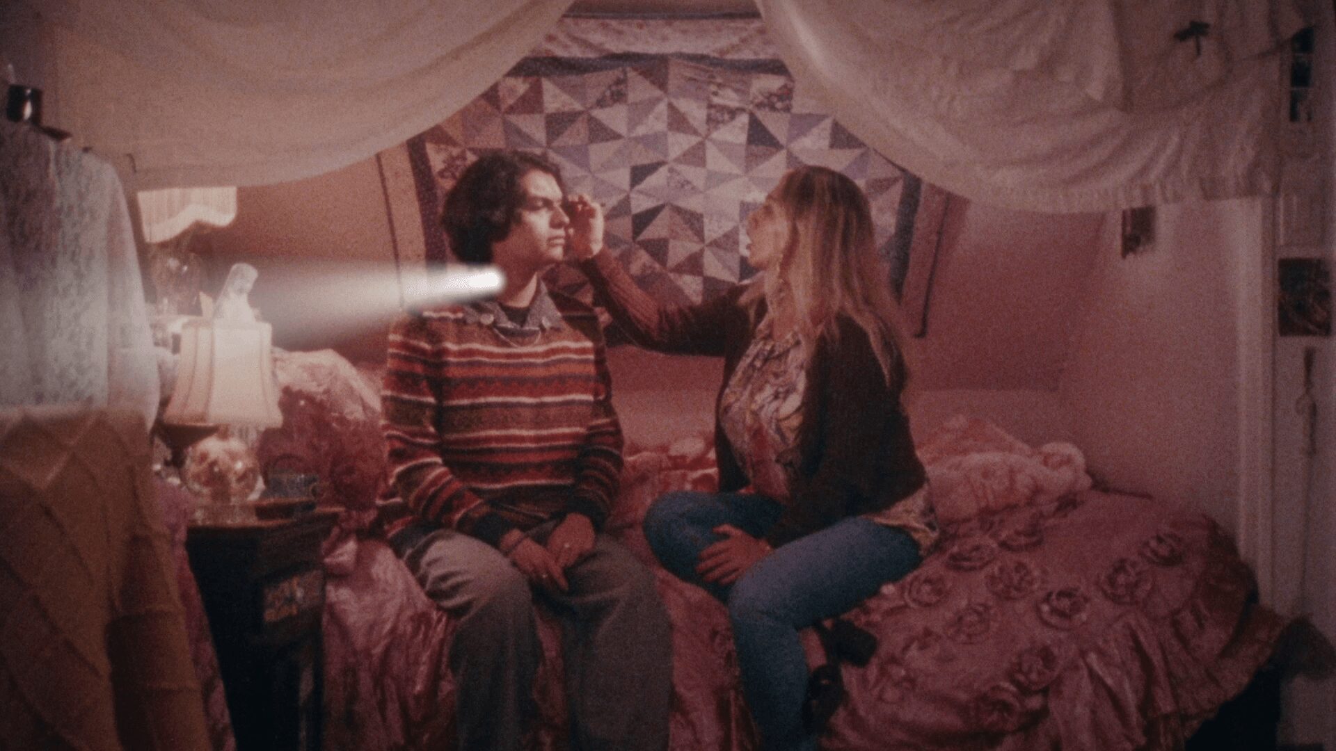 Two women sit on a bed in a cozy room, one touching the other's shoulder, with a quilted wall hanging behind them.