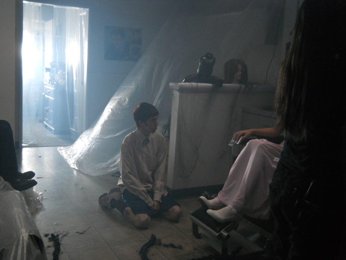 Person sitting on a chair with legs crossed, another person kneeling on the floor, light coming through a window, and a curtain.