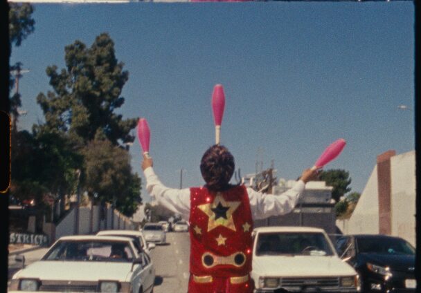 Clown with pink juggling clubs standing in a parking lot with cars and trees in the background.