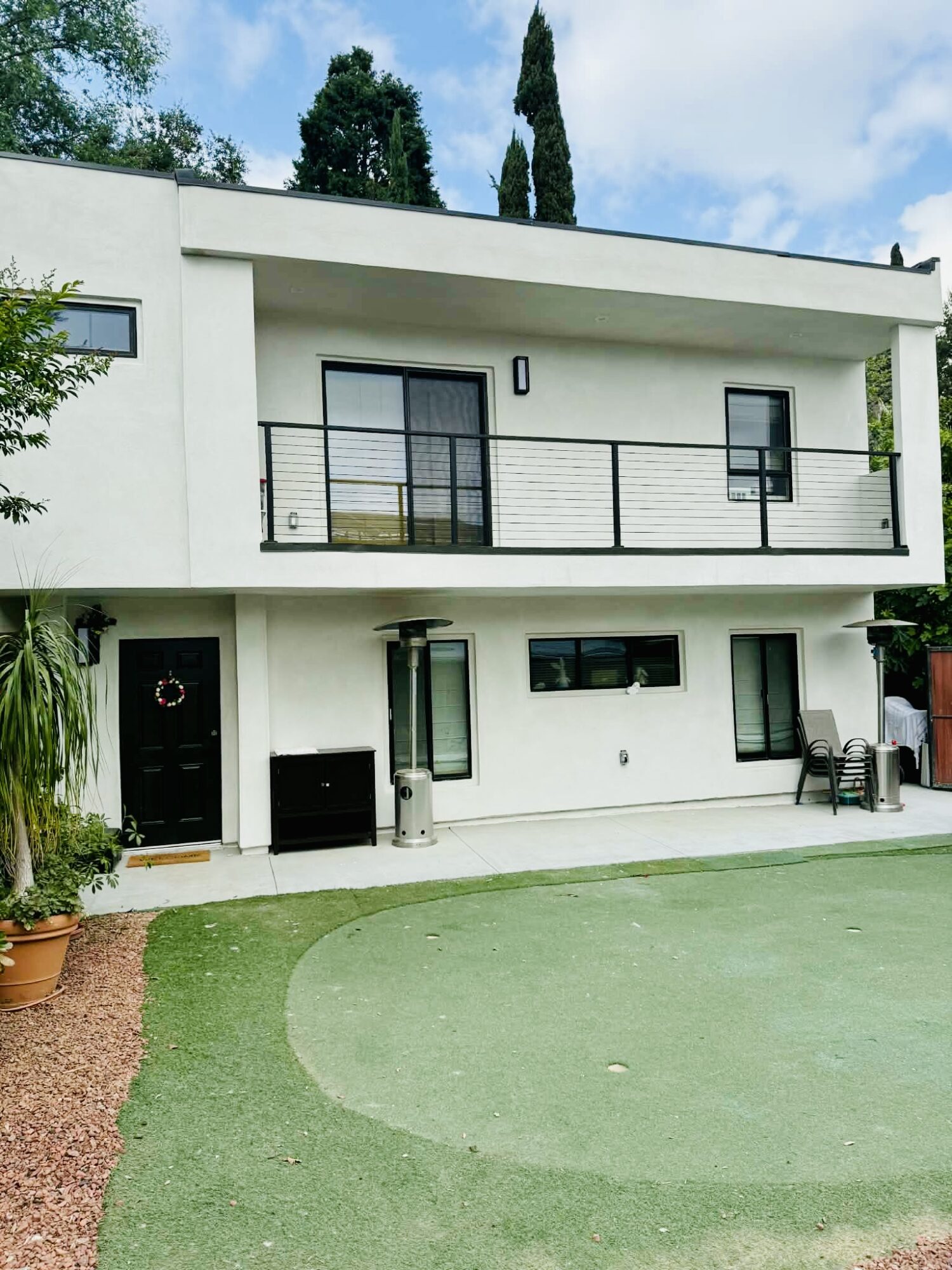 Modern two-story house with balcony, black railing, and white exterior, surrounded by green lawn and trees.