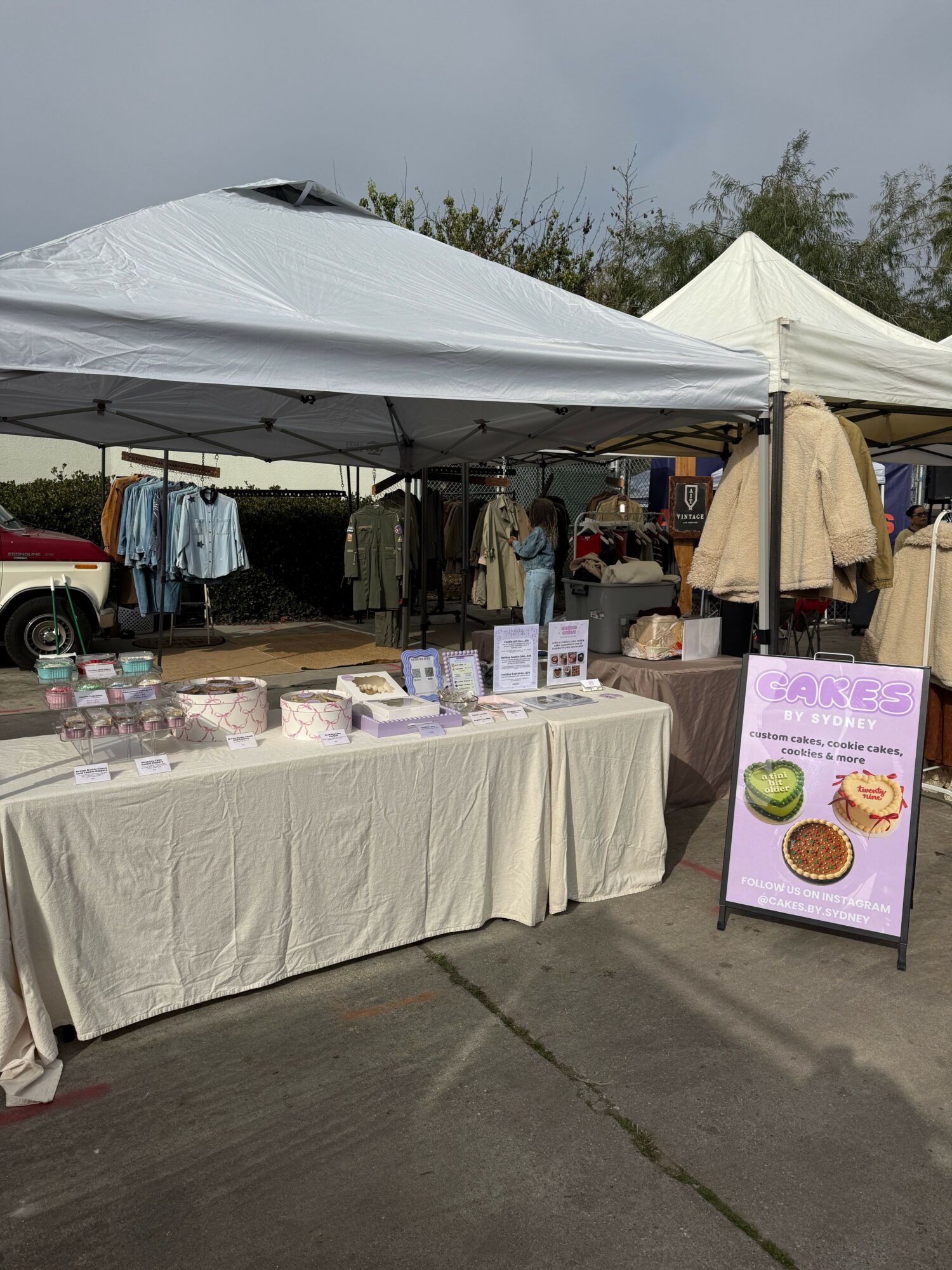 Outdoor market stall with clothing and baked goods under tents, cloudy sky, and trees in background.