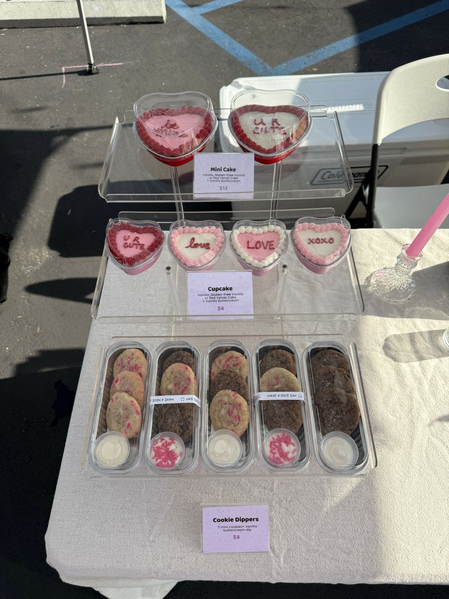 Display of heart-shaped and round cookies with pink and red icing on a table outdoors, with a white chair and shadow nearby.