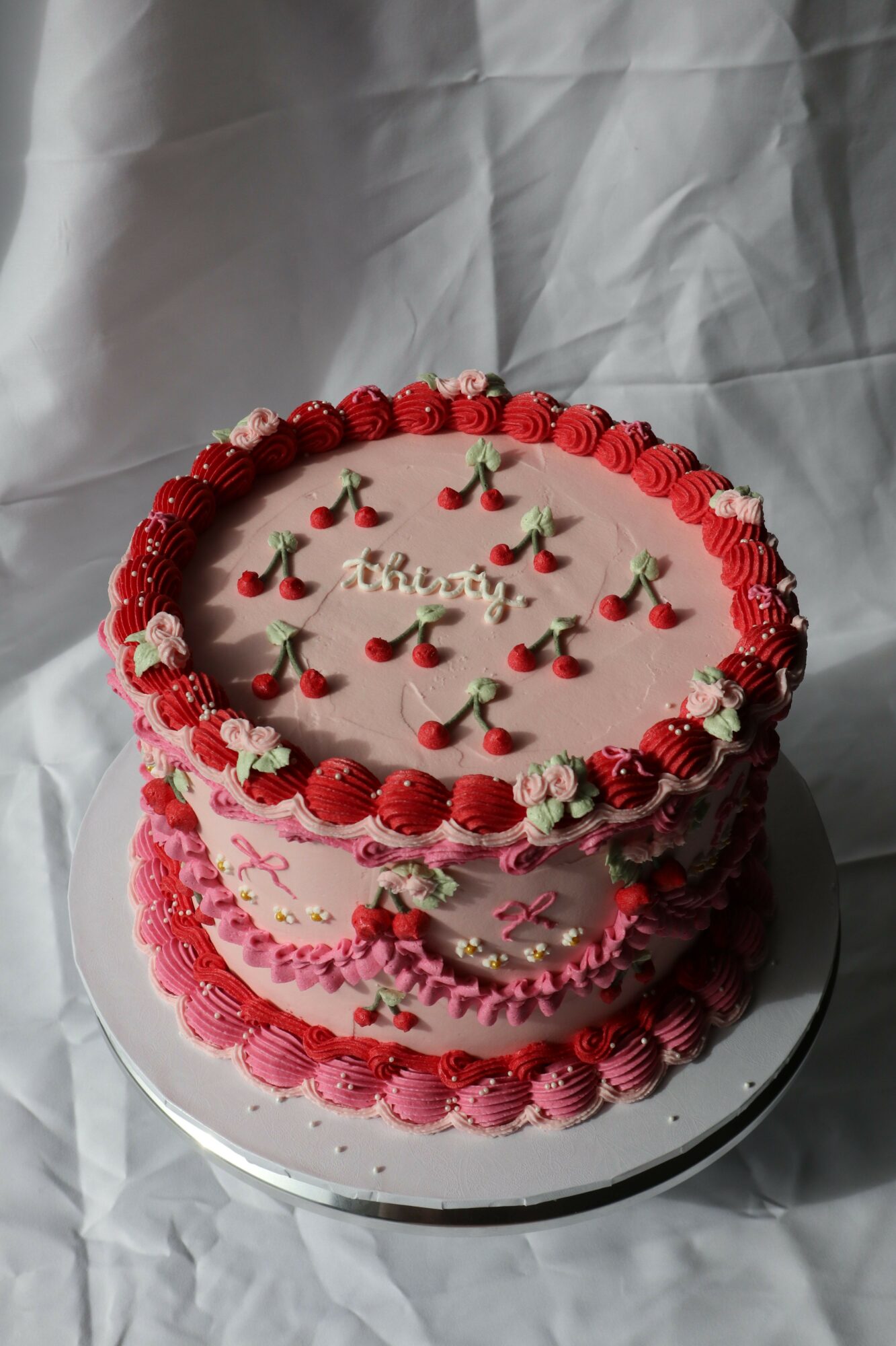 Two-tiered birthday cake decorated with red and white icing, strawberries, and small flowers, on a white surface.