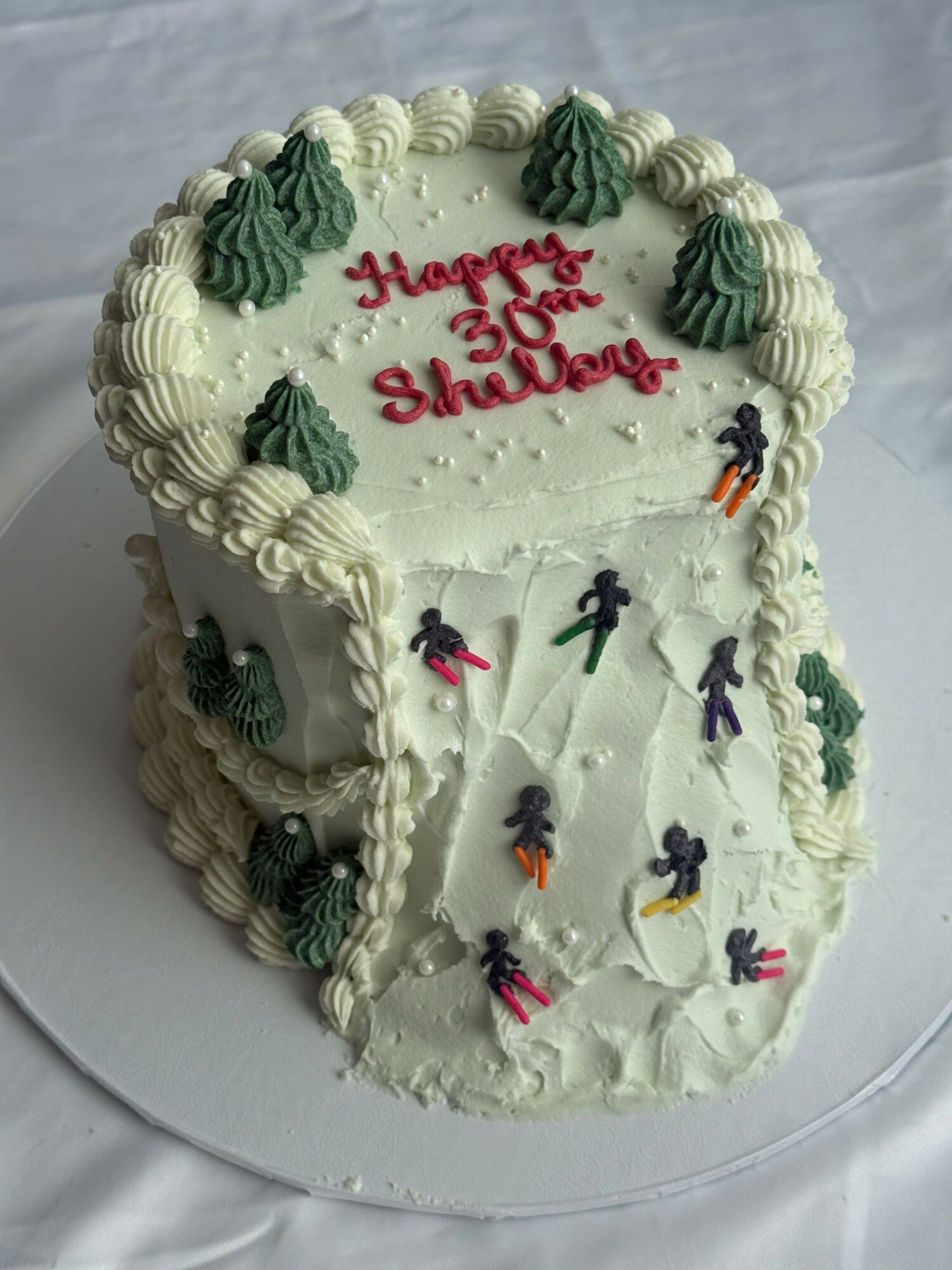 Cake with white frosting, decorated with green trees, ice skaters, and red writing for a 50th birthday celebration.