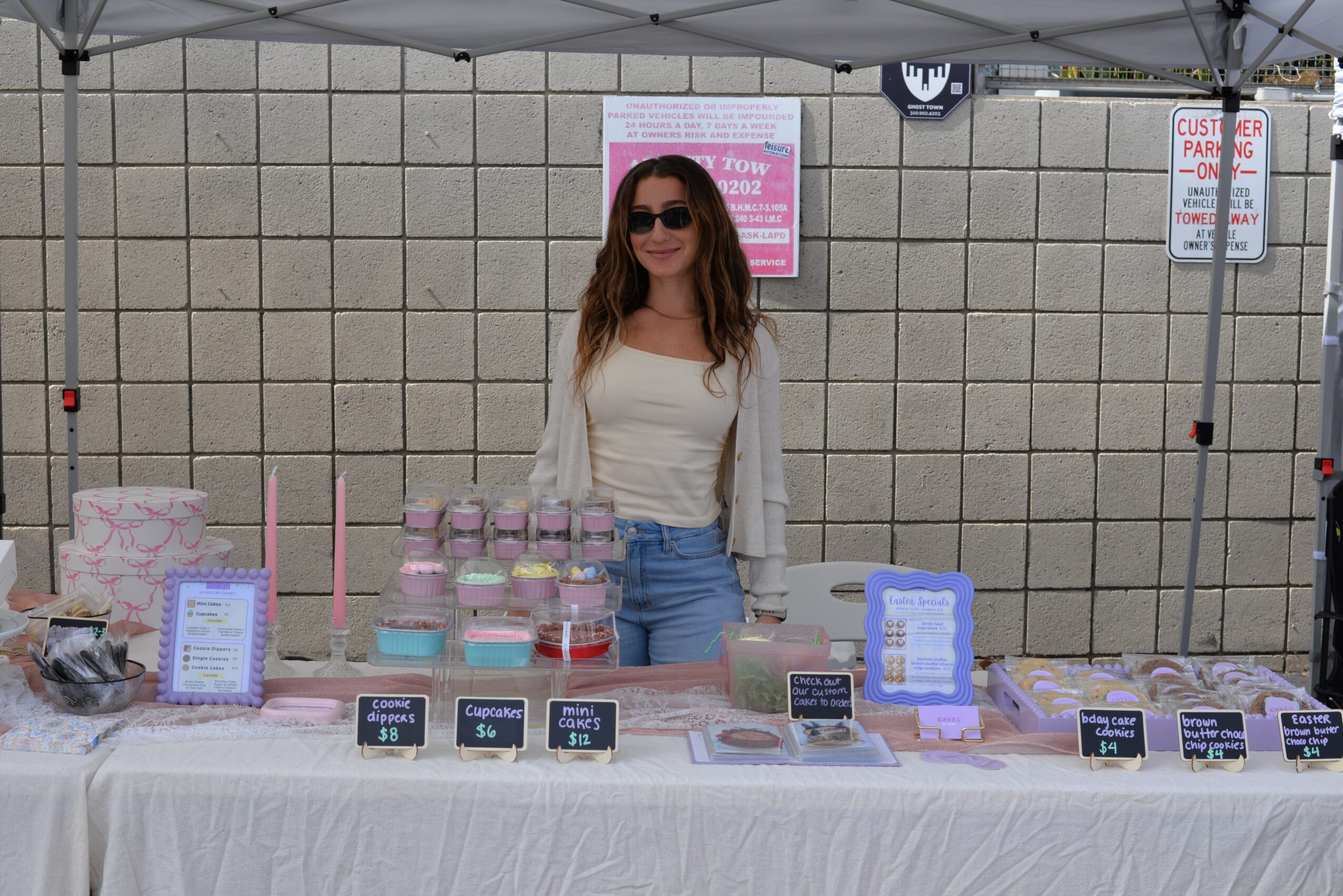 Woman with sunglasses standing behind a table with pink and white products at an outdoor market stall.