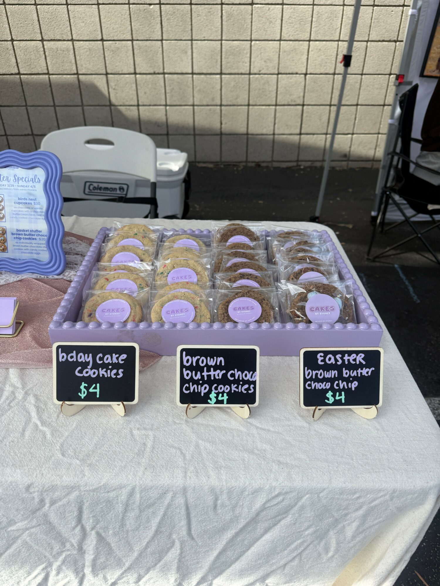 Table with three trays of cookies labeled for Easter, brown butter, and bday cake, with small chalkboard signs displaying prices.