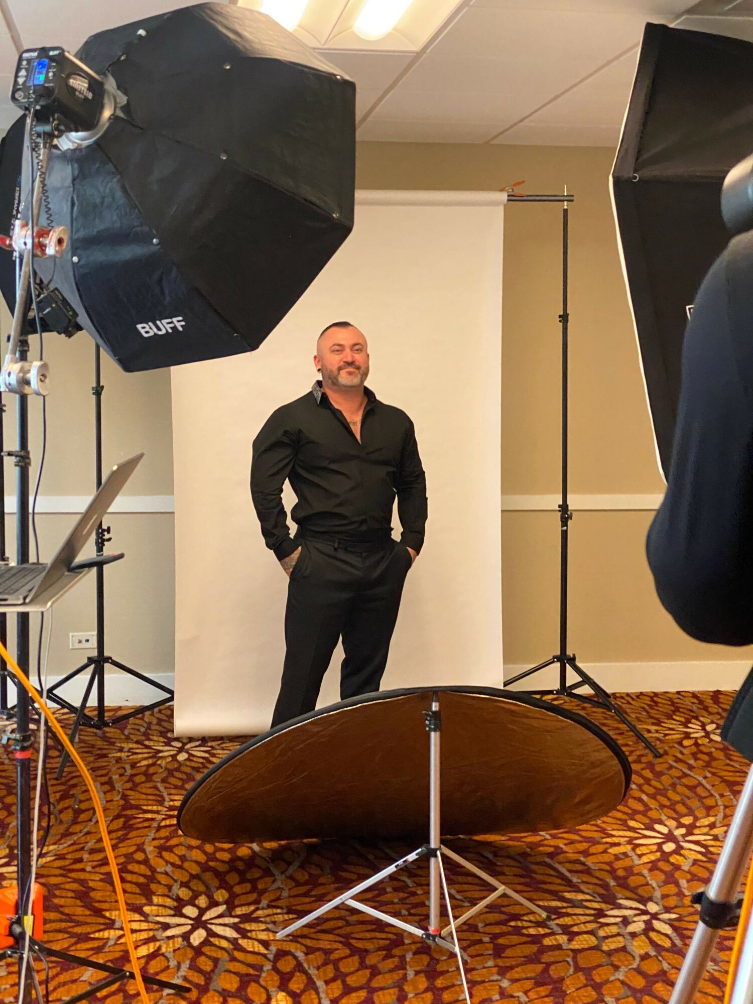 Man in black shirt standing in front of a white backdrop, surrounded by photography equipment, in a room with patterned carpet.