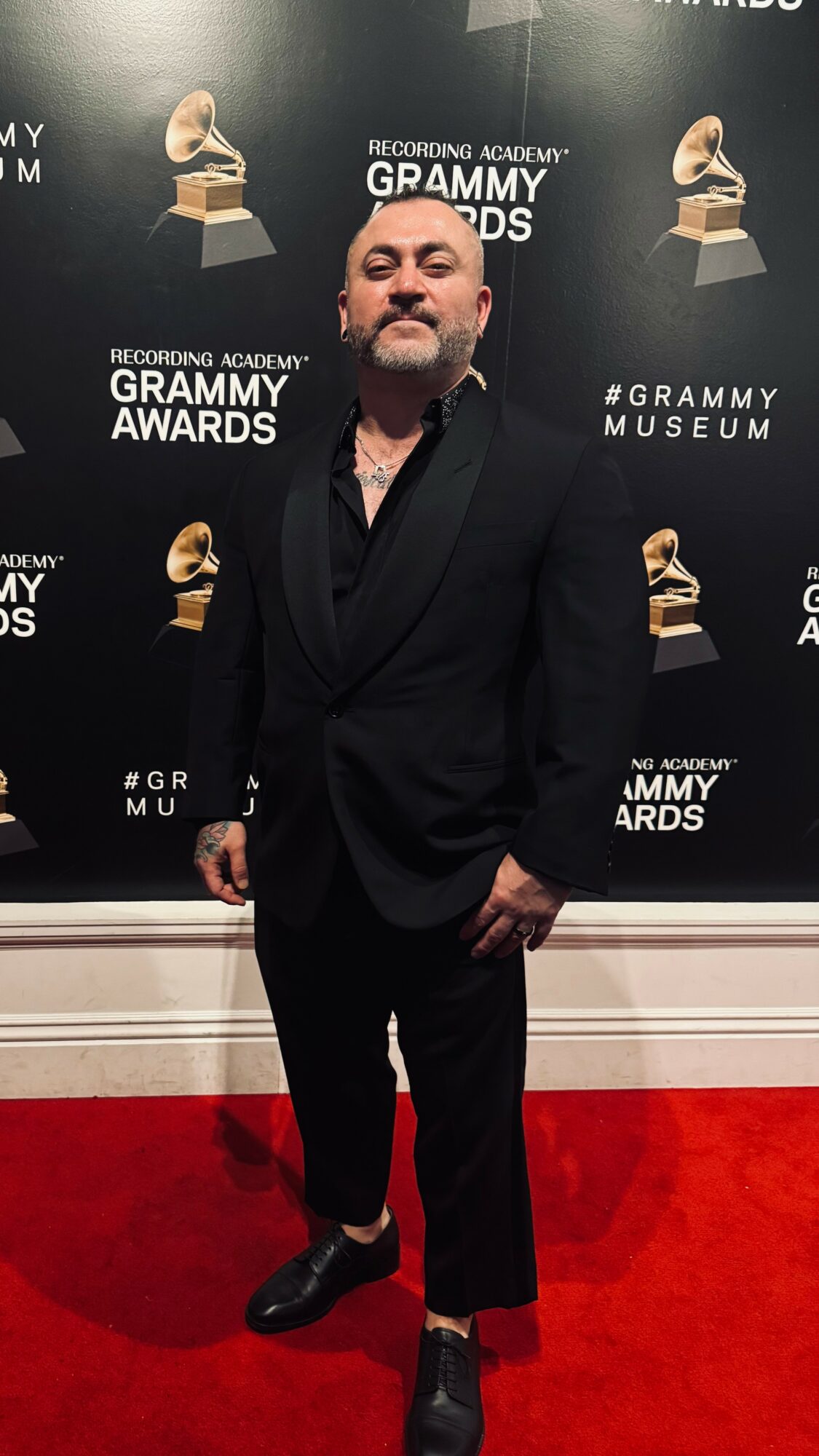 Man in black suit standing on red carpet at Grammy Awards event, backdrop with Grammy Museum logos.