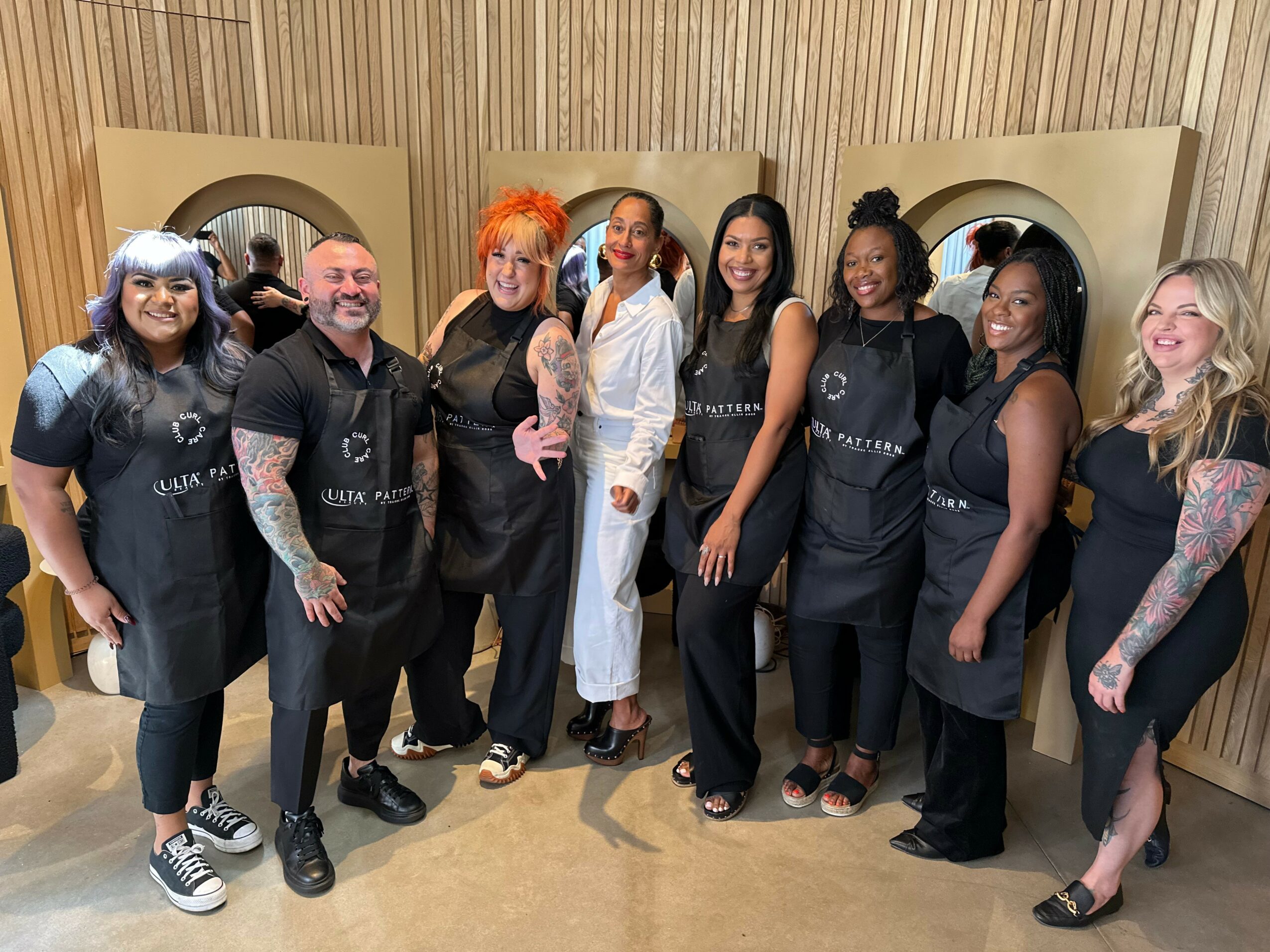 Group of seven people standing indoors, smiling, some with tattoos, wearing black uniforms or outfits, wooden wall background.