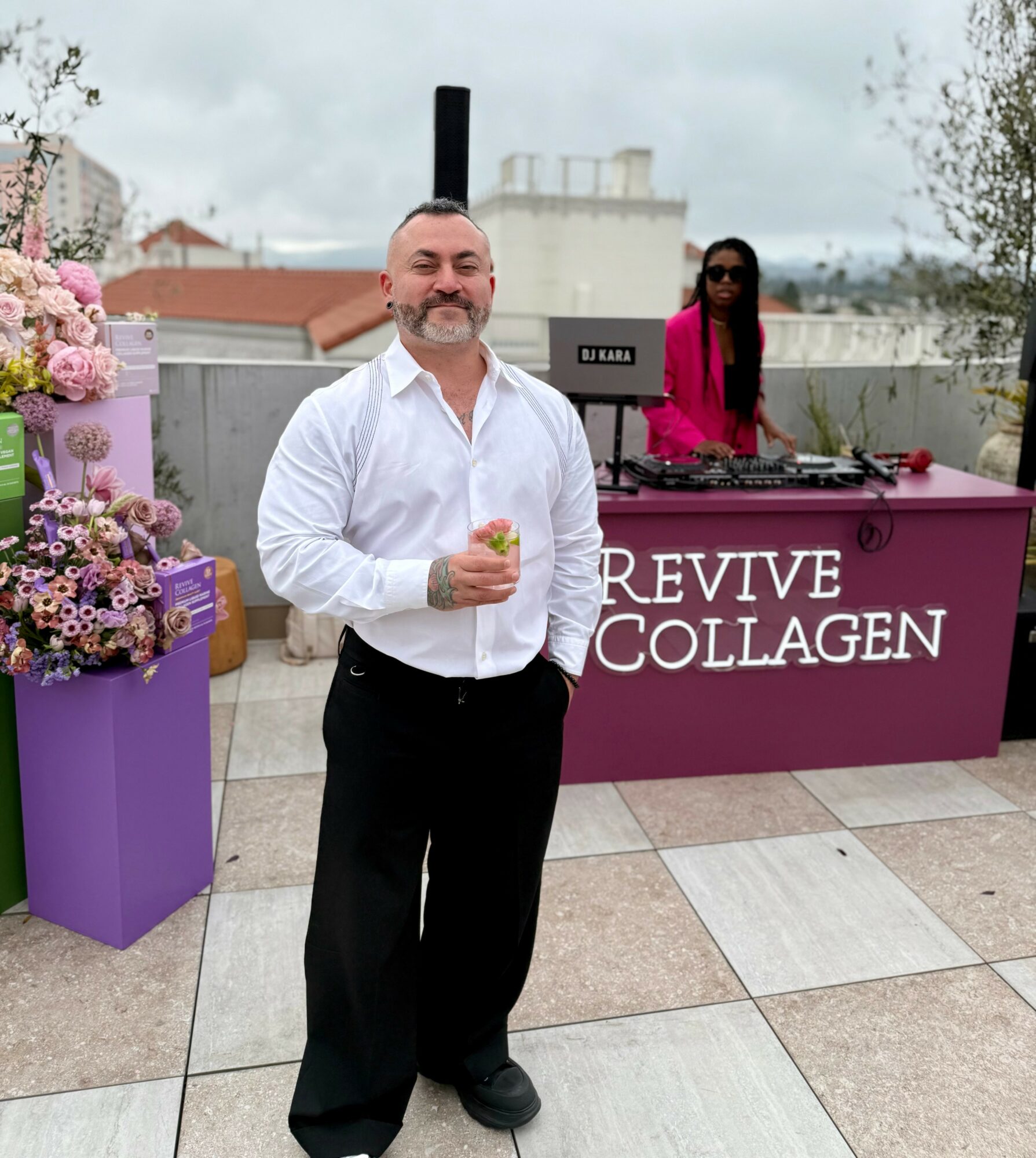 Man in white shirt holding a drink on rooftop with DJ and city view in background.