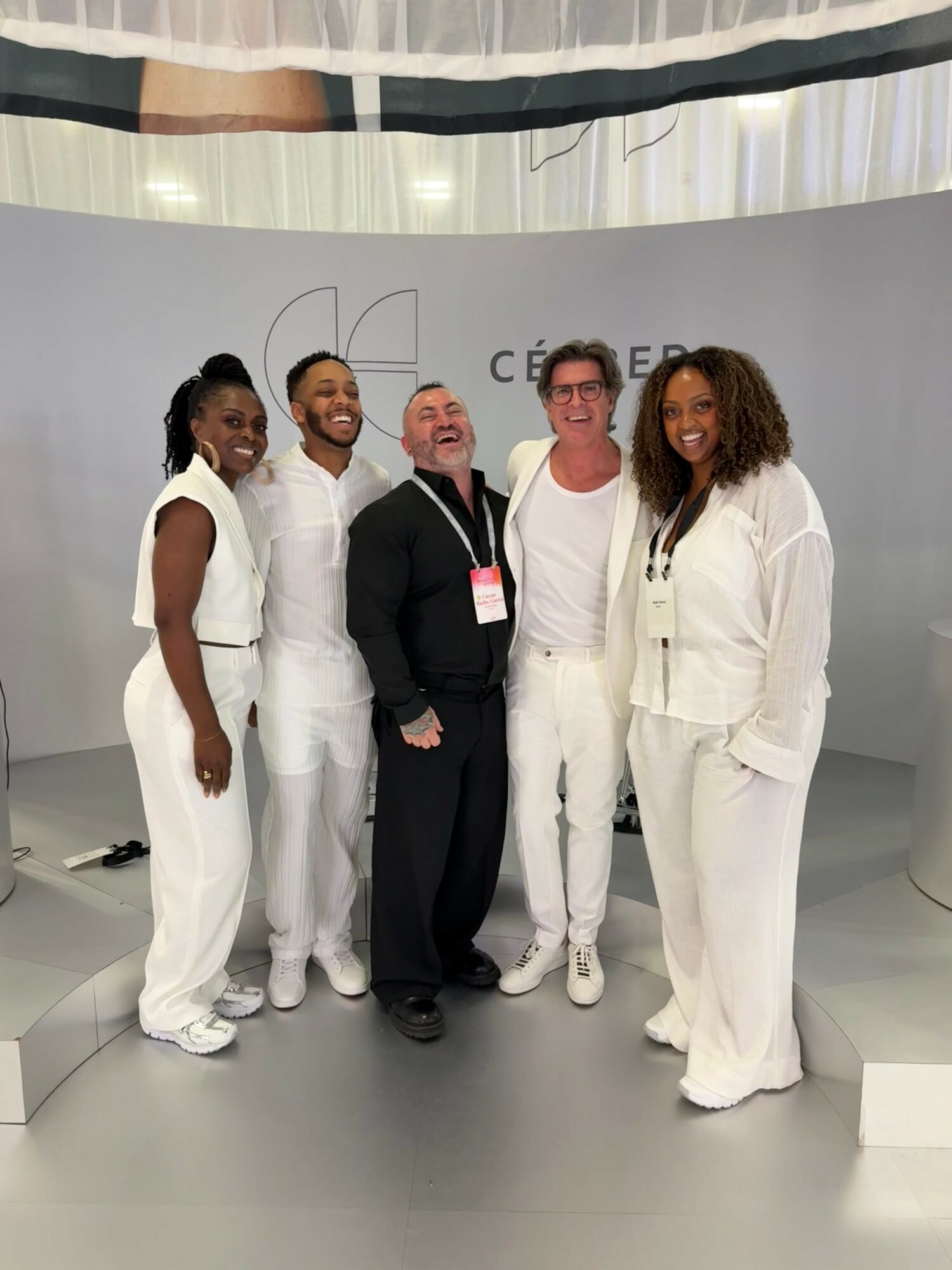 Five people standing together on stage, smiling, with a white backdrop and curtains above.
