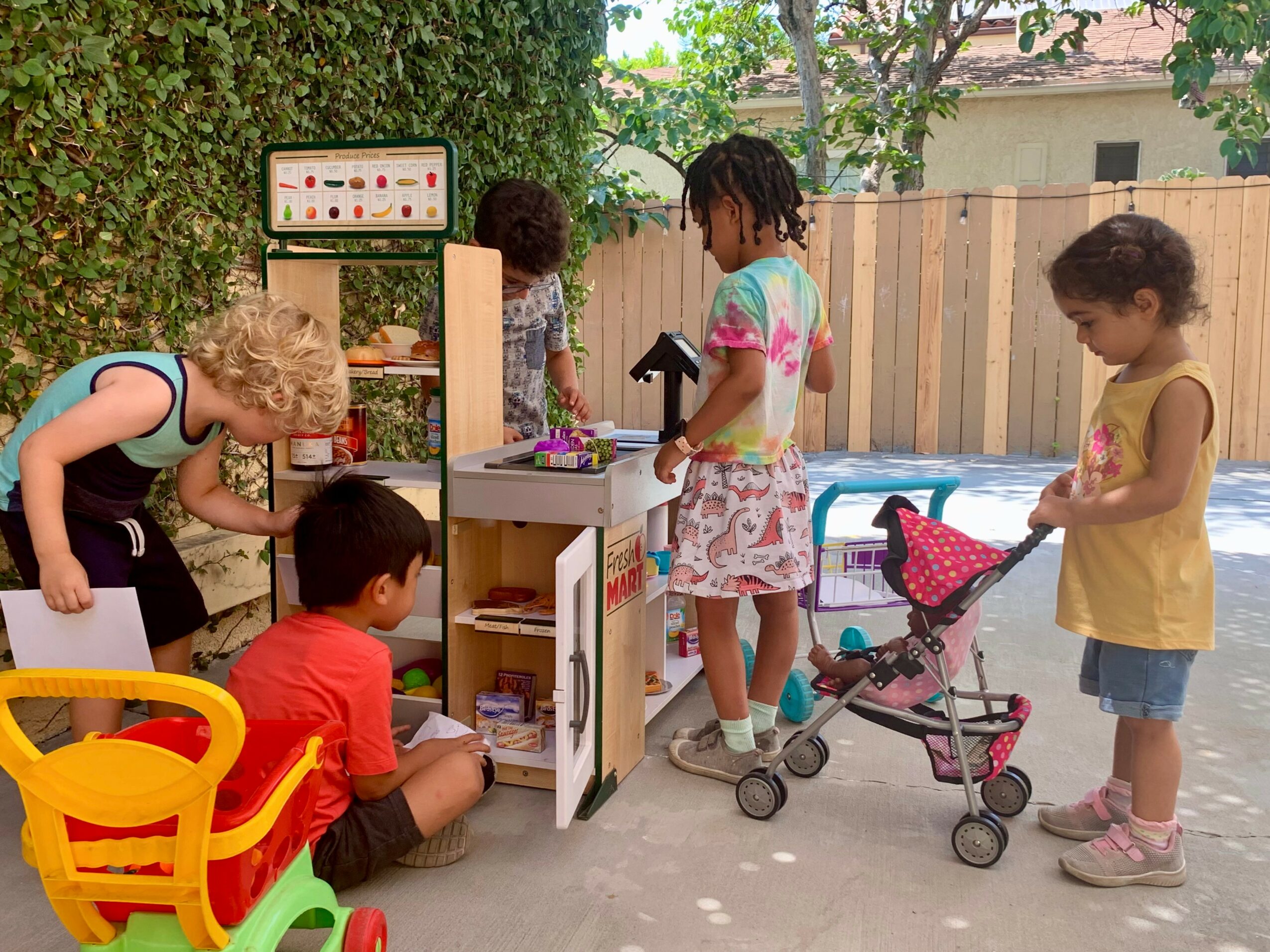 Group of children playing outdoors with toys and a woman supervising, in a backyard with trees and a fence.