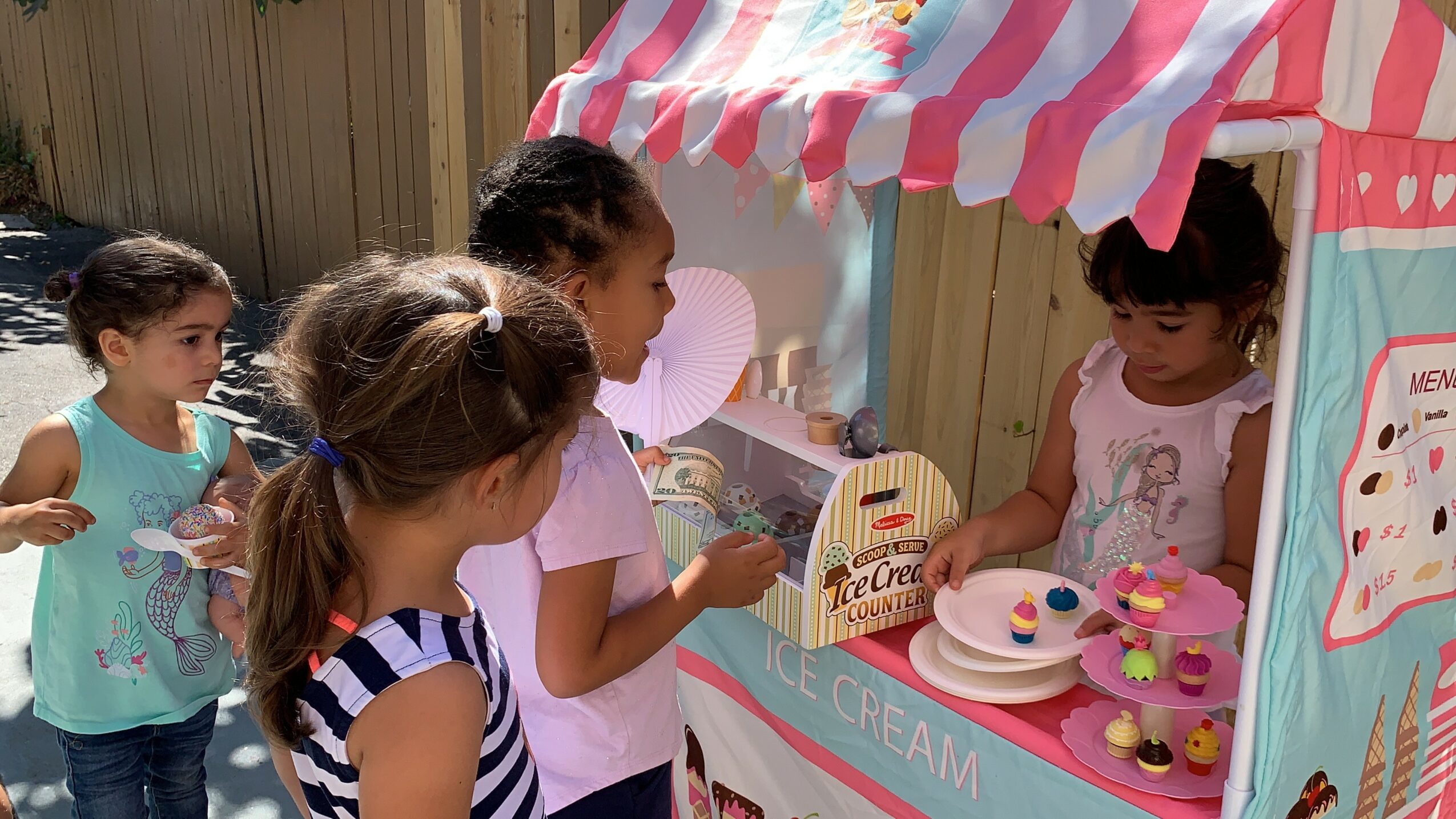 Four children at a pink and white ice cream stand, one girl serving ice cream, others waiting.