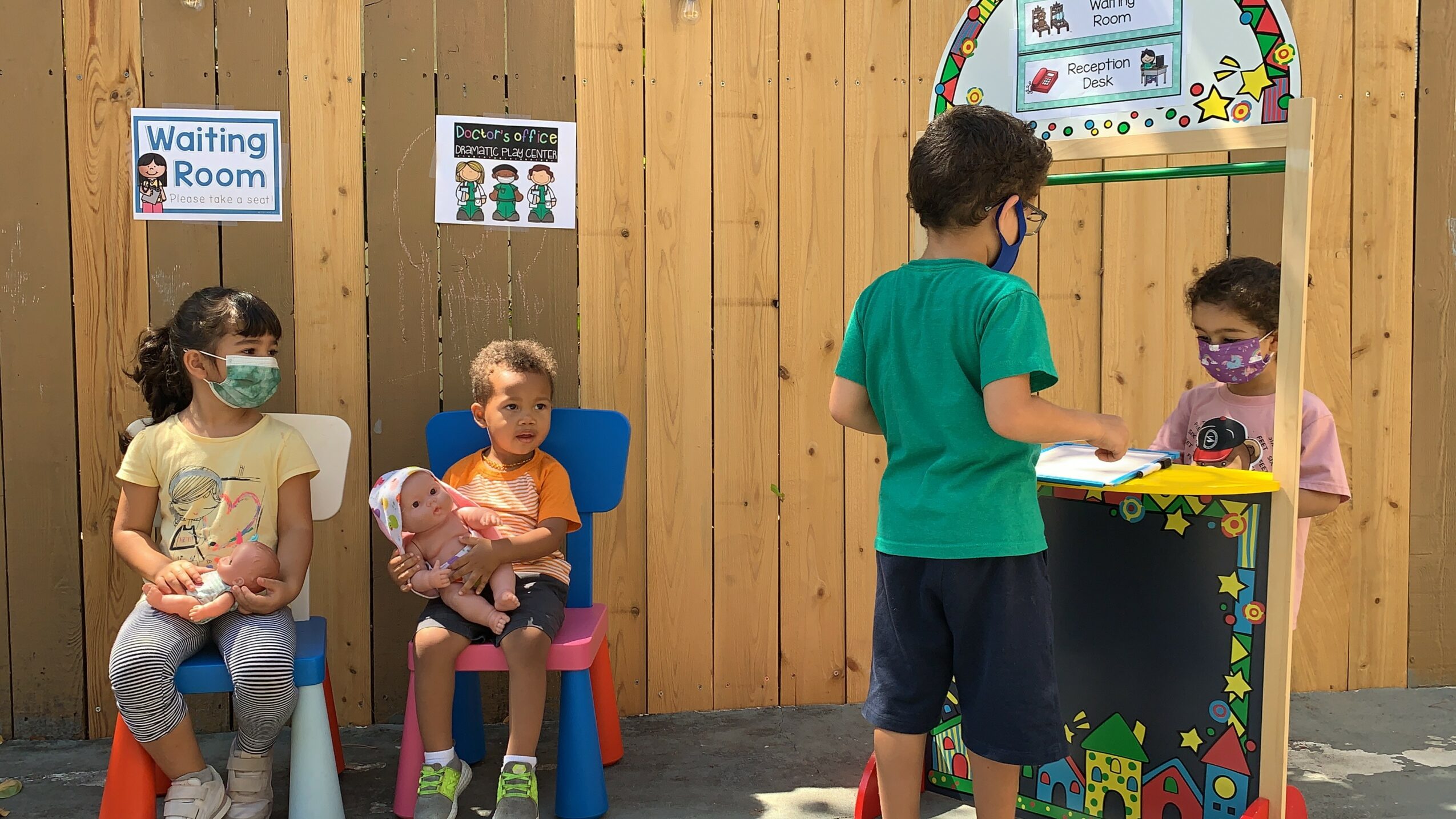 Three children and a woman at a play area with wooden walls, two children sitting, one standing, woman interacting at a booth.