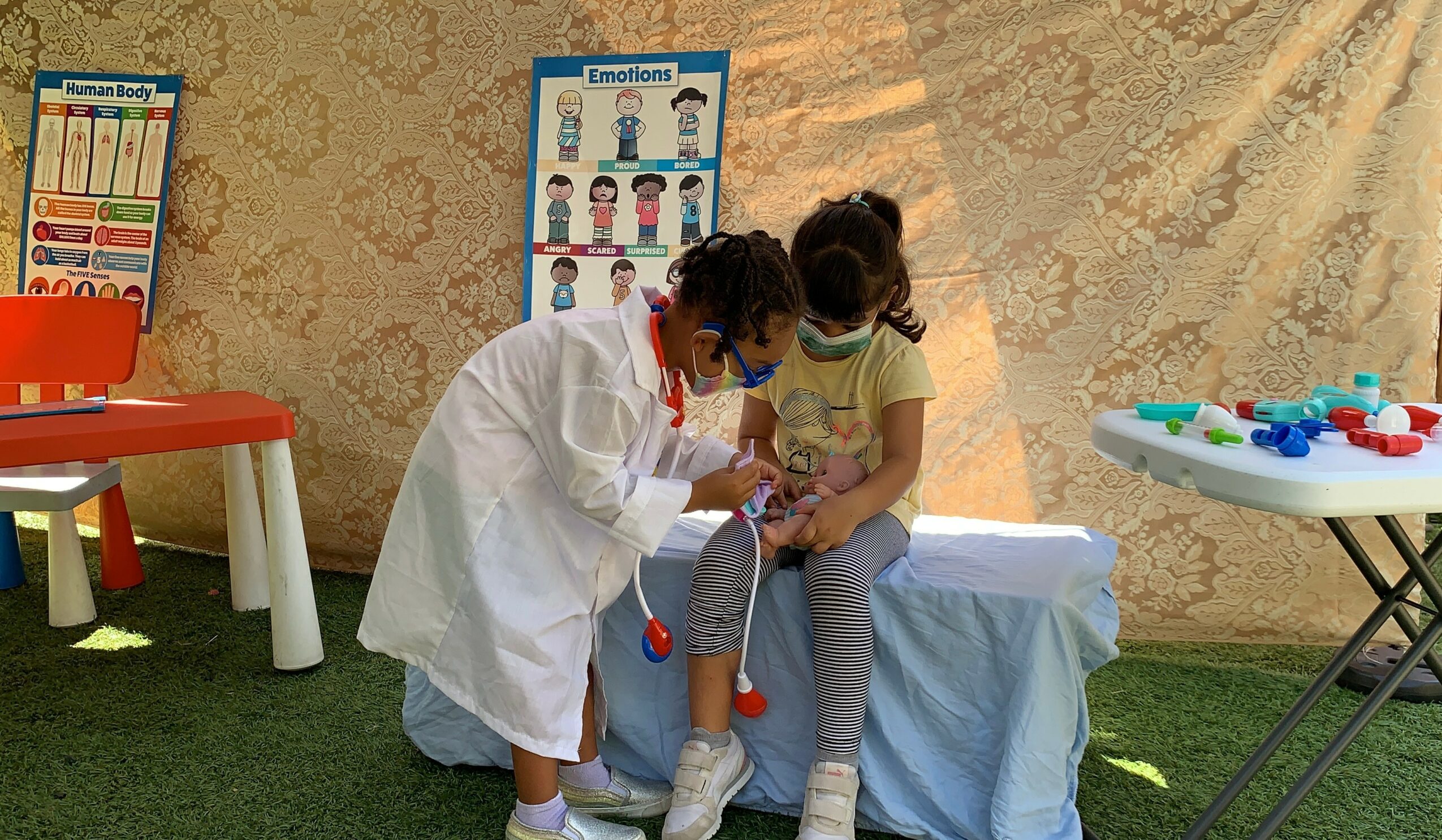 Child in a white coat and mask interacts with a seated girl holding a medical device outdoors.