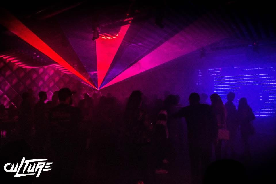 Dark nightclub scene with colorful laser lights and silhouettes of people dancing and socializing.