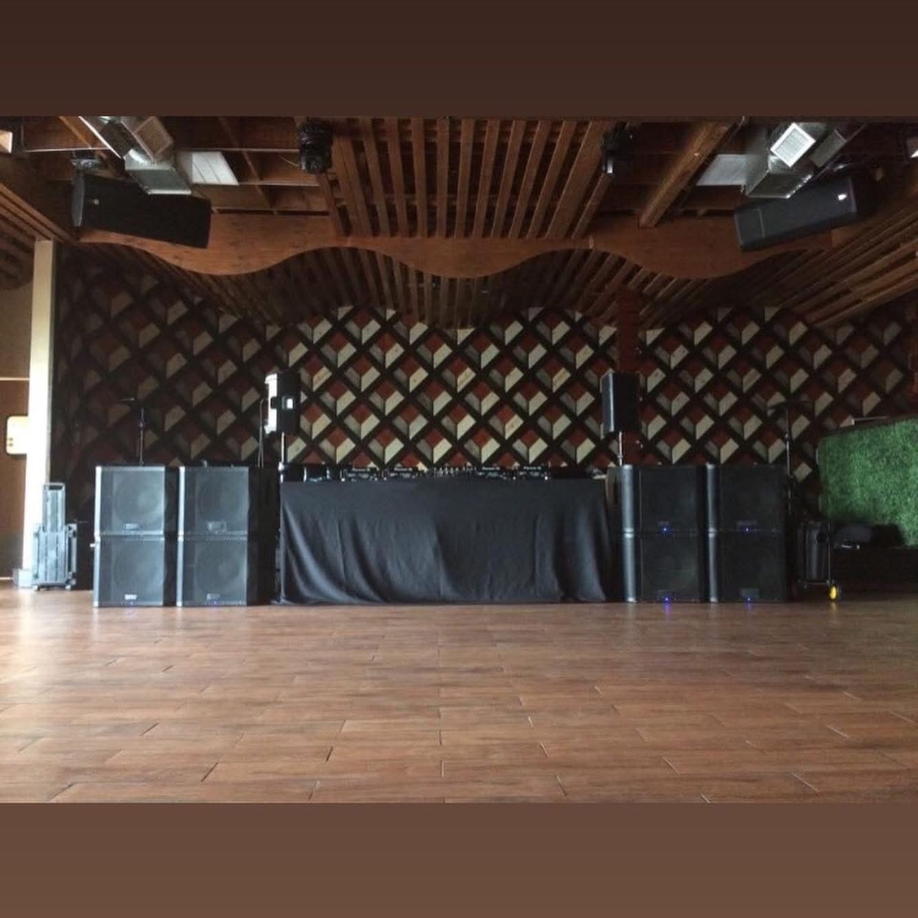 DJ booth with speakers and equipment on a stage, wooden ceiling, patterned wall, and wooden floor.