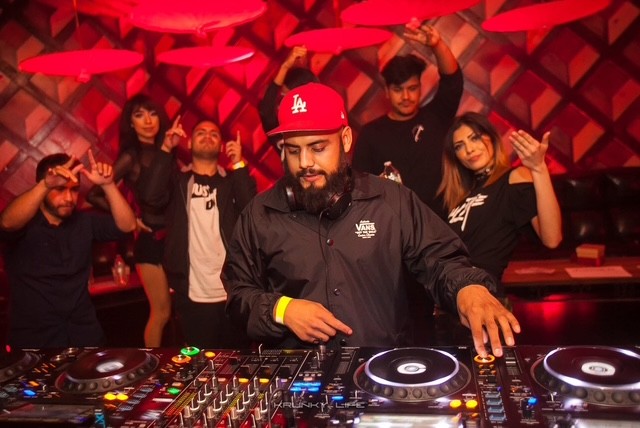 DJ with beard wearing red cap and black jacket, mixing music at a club with five people dancing behind him.
