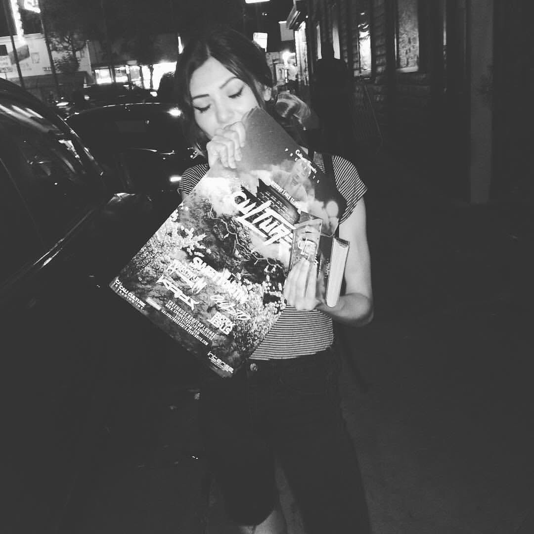 Young woman reading a magazine on a city street at night, with parked cars and buildings in background.