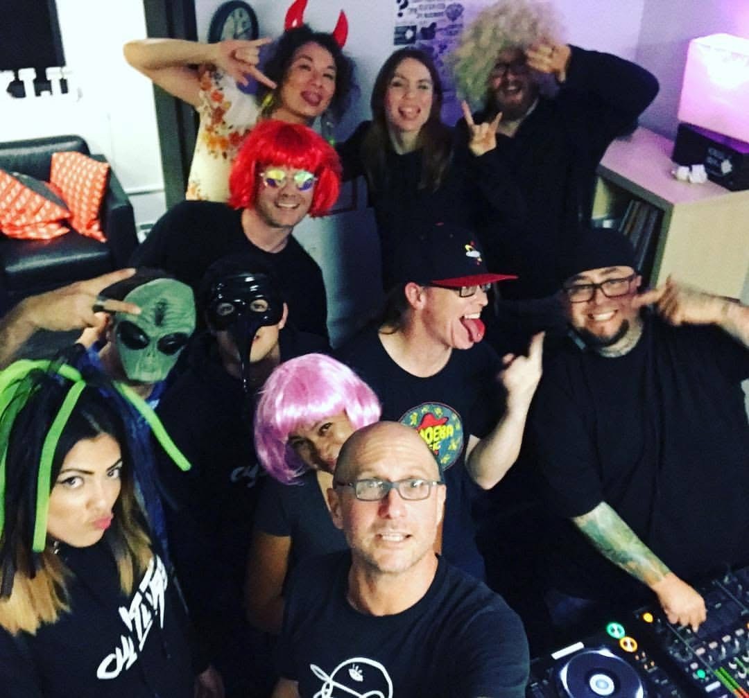 Group of people in costumes and wigs, some wearing glasses, posing indoors with DJ equipment and a TV in background.