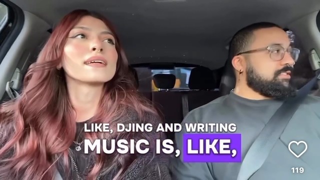A woman with long red hair and a man with glasses and a beard sit in a car, talking.