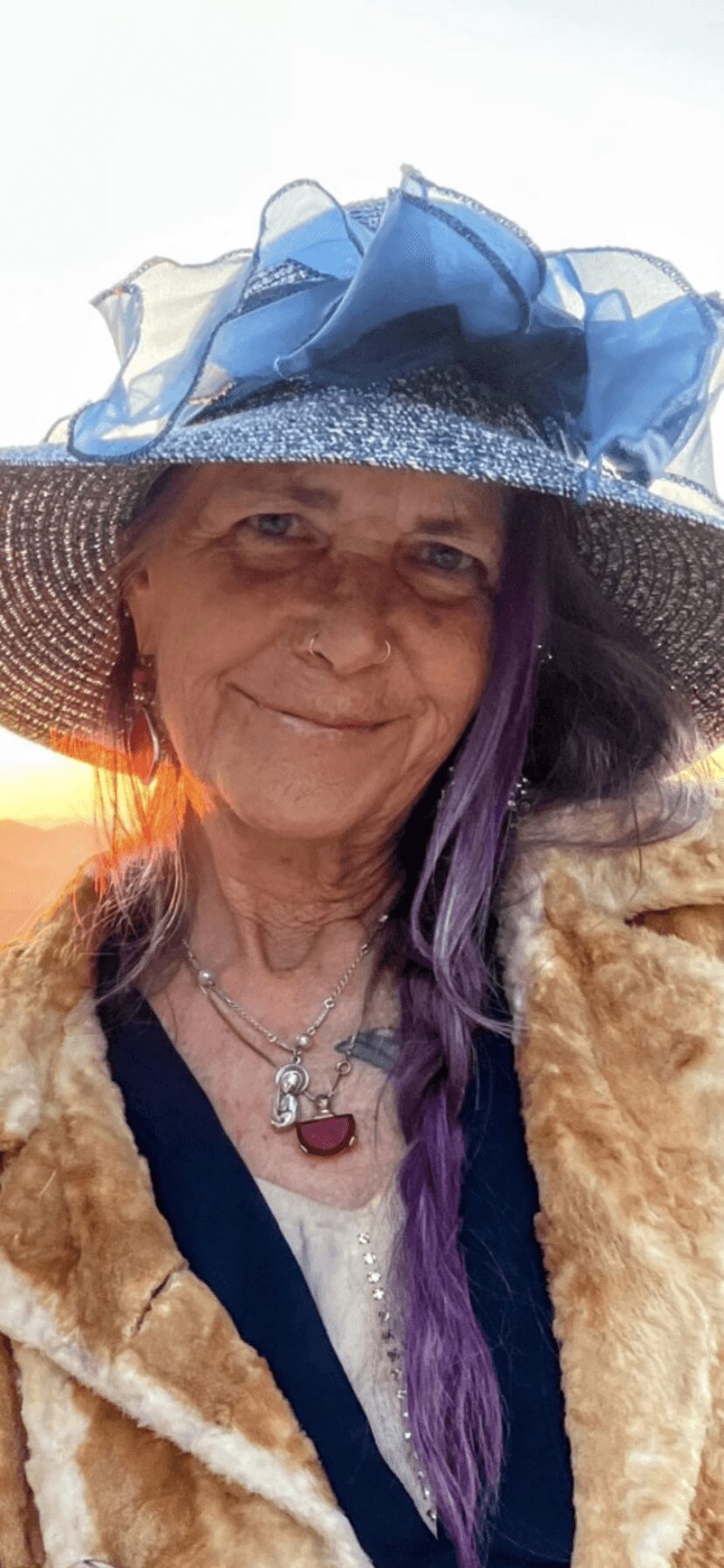 Woman wearing a large hat with blue decorations, smiling, with purple hair and layered clothing.