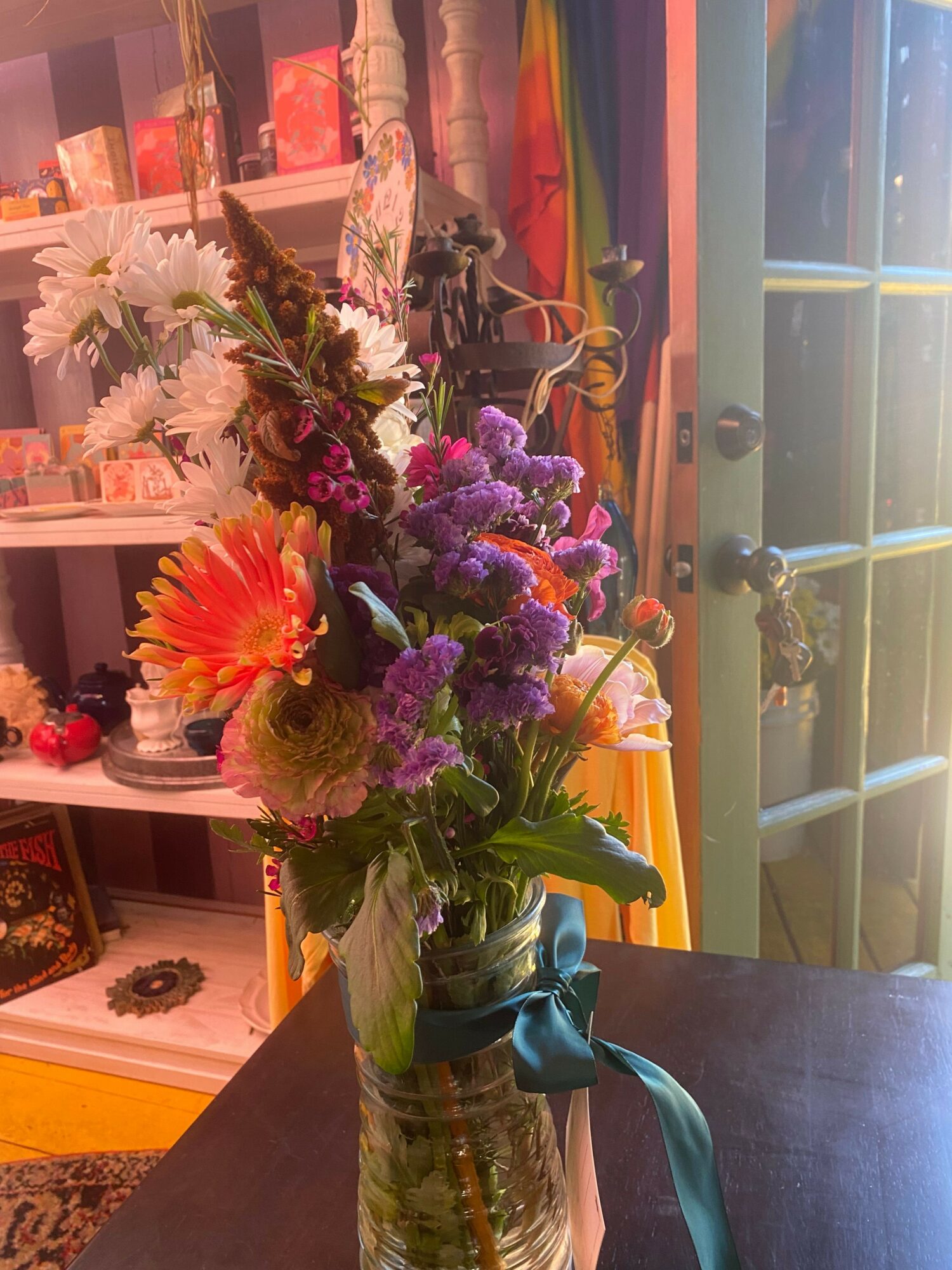 Colorful mixed flower bouquet in a glass vase on a dark table, with a window and colorful curtains in the background.