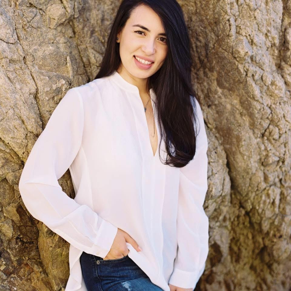 Young woman with long dark hair smiling, wearing a white blouse and blue jeans, standing outdoors against a rocky background.