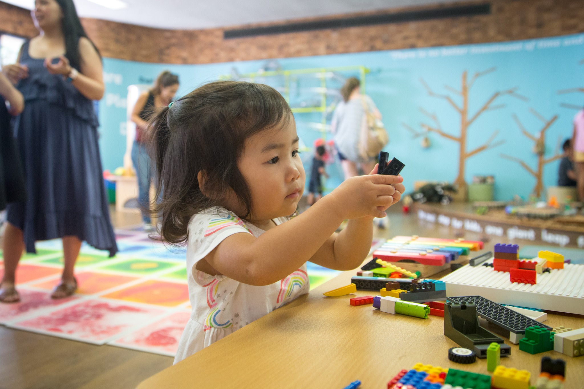 Young girl playing with colorful building blocks at a table indoors, with other children and adults in the background.