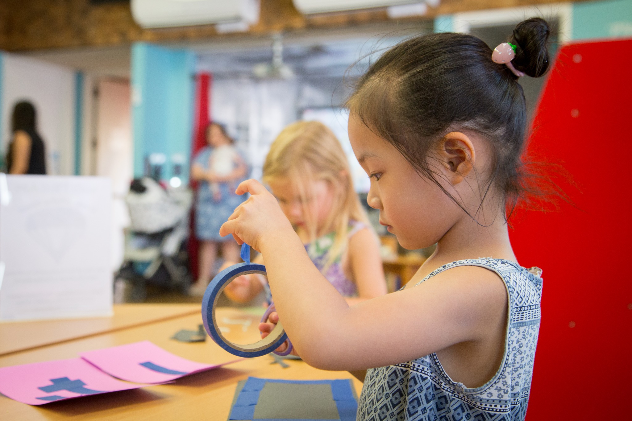 Young girl with dark hair in a bun, wearing a sleeveless top, looks at a magnifying glass at a table.