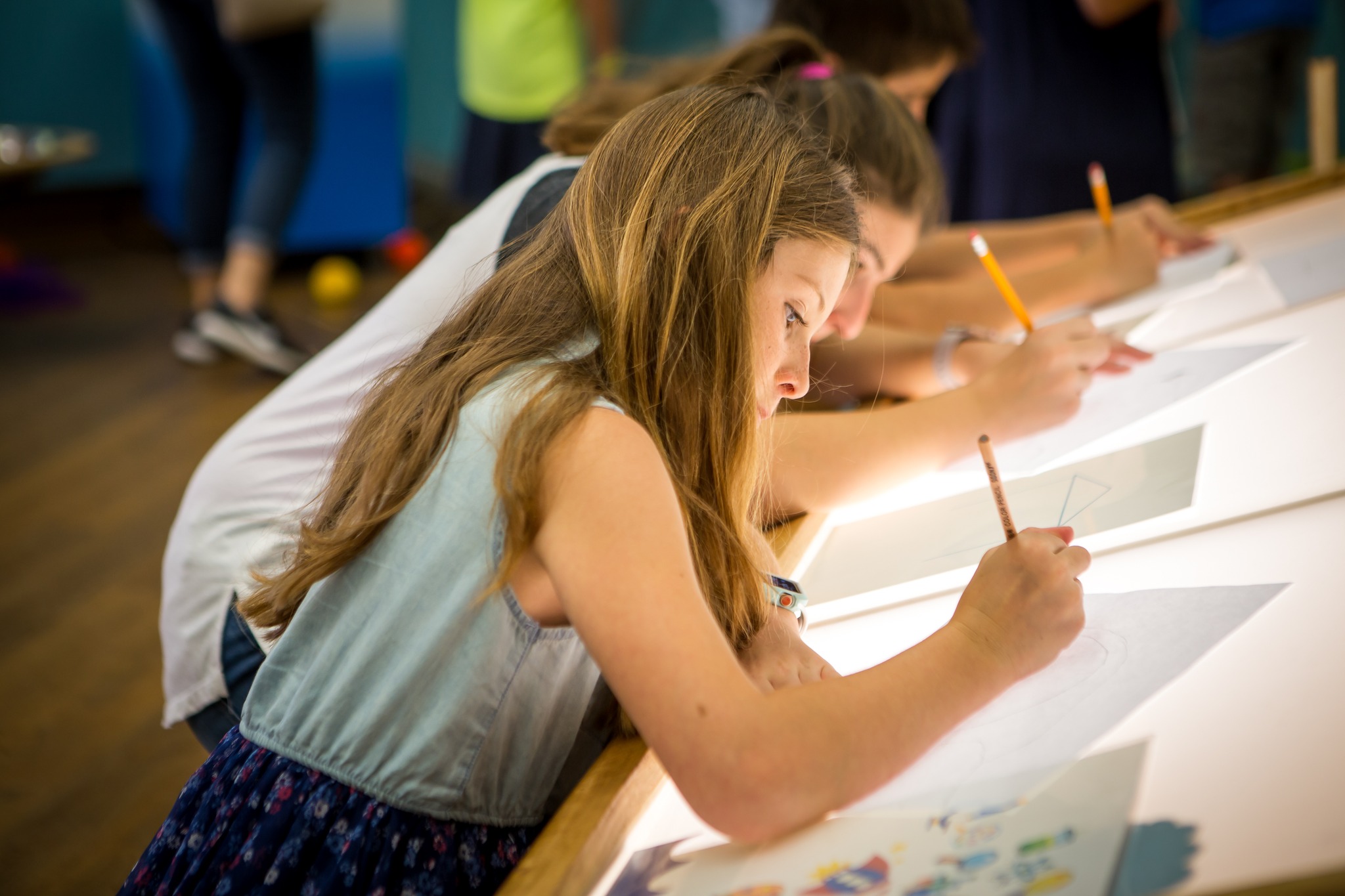 Girl with long hair writing on paper at a table, surrounded by others also writing.