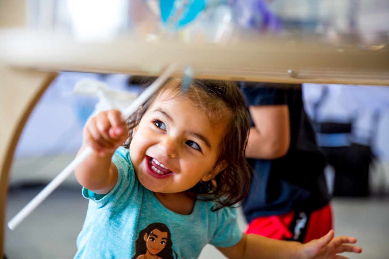 Young girl smiling and reaching under a table, wearing a blue shirt with a cartoon character.